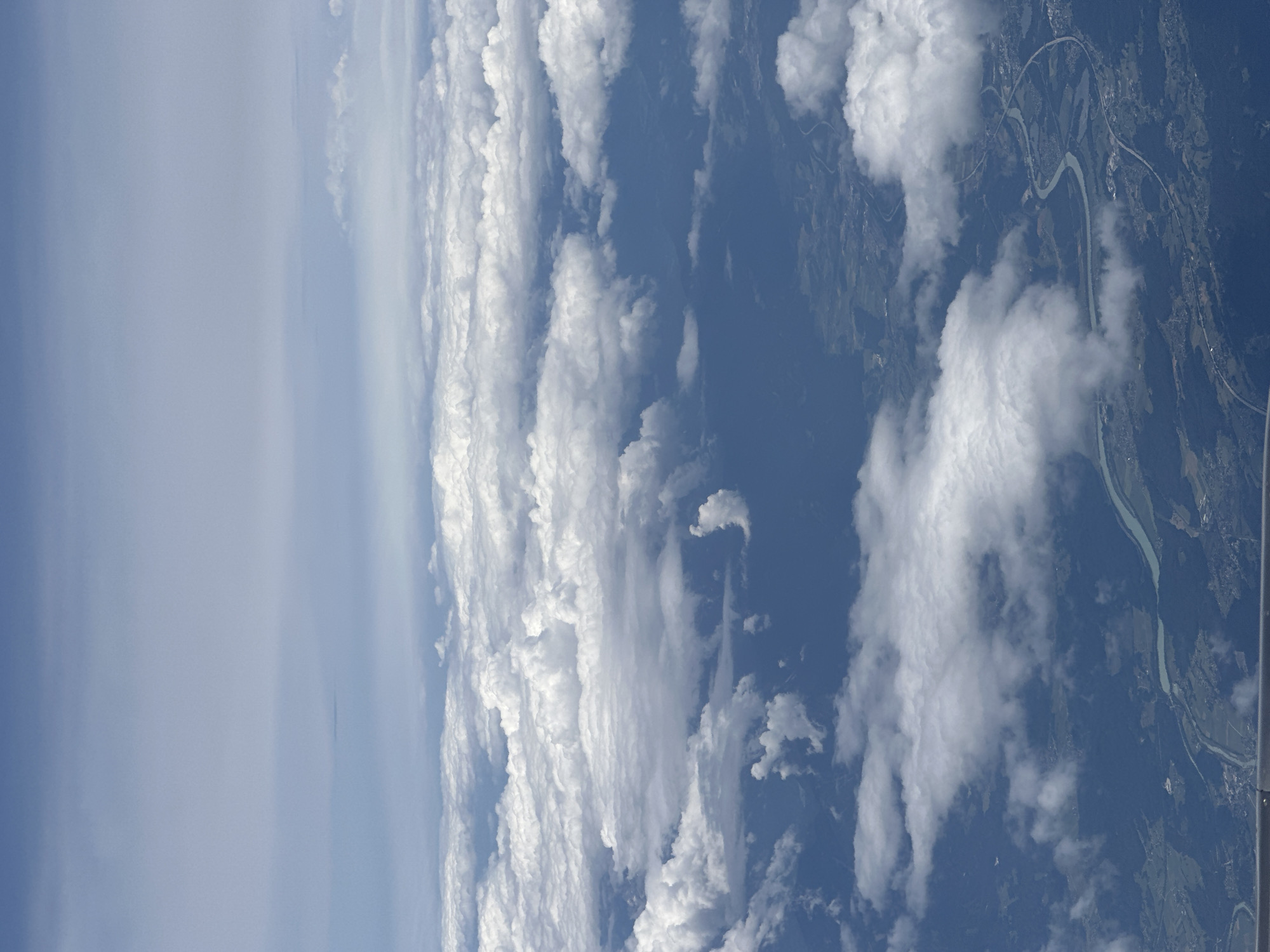 An aerial view captures a landscape from an airplane, dominated by a vast expanse of fluffy white clouds. Below the clouds, the earth is visible, showing a winding river with a turquoise hue. Along the riverbanks and surrounding areas, lush green terrain is interspersed with dark forests and scattered human settlements, indicating a rural or suburban environment. A road or highway network is visible, curving alongside the river and through the landscape, with some infrastructure and buildings visible. The sky above is a pale blue, fading into a lighter hue towards the horizon, with faint haze or distant clouds. There are no people or specific actions depicted in the image; it is a static, environmental scene. The crops suggest the image might be related to Glantschach, Austria, given the geographical features like the river and topography.