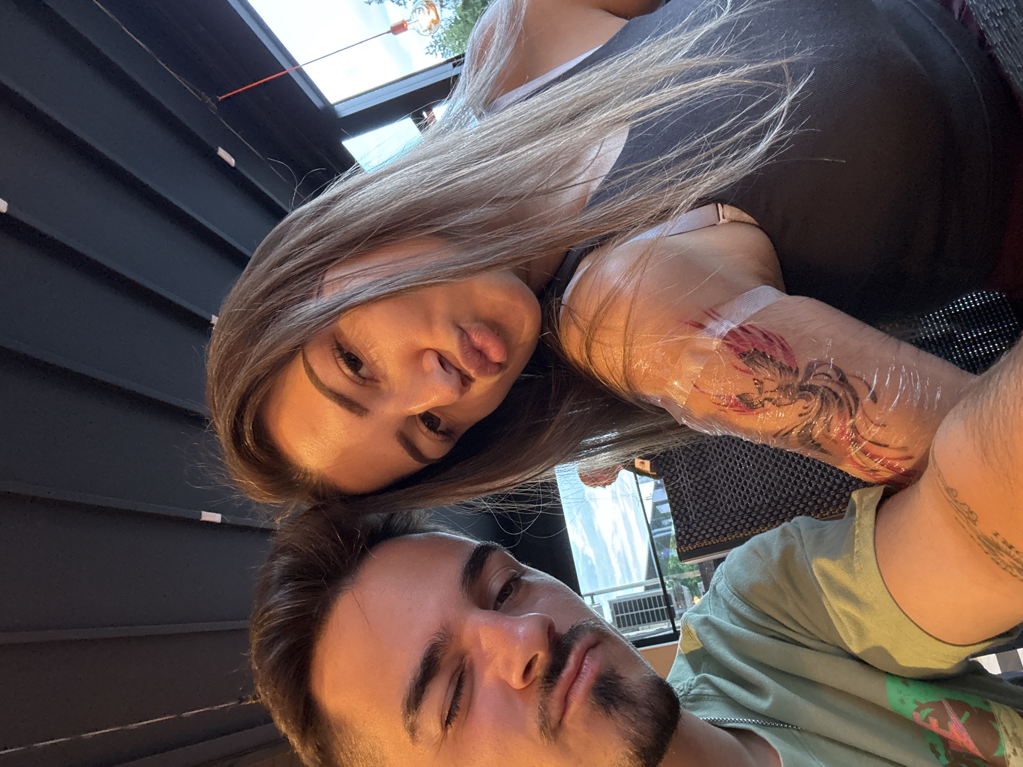 This close-up selfie, captured in Sofia, Bulgaria, features two individuals: a woman and a man. The image is taken from a slightly low angle, looking upwards.
The woman is in the upper part of the frame, looking directly at the camera with a playful "duck face" expression, her lips puckered. She has long, light brown hair that cascades over her shoulder. She is wearing a dark top, with a light-colored bra strap visible on her right shoulder. A prominent feature is the fresh tattoo on her right forearm, which is covered with clear protective plastic film. The tattoo appears to be a colorful, intricate design, possibly depicting a creature or elaborate pattern. Another, less distinct tattoo is visible on her upper right arm.
The man, who appears to be taking the photo, is positioned in the lower part of the frame. He has short, dark brown hair and a neatly groomed beard and mustache. He is looking towards the camera with a relaxed expression, his eyes slightly narrowed or partially closed. He is wearing a light green t-shirt with a design partially visible on the front.
The setting appears to be an indoor or semi-outdoor space, possibly a cafe or restaurant. The background on the left features a dark, slatted wall or paneling. Above this, a skylight or window reveals a bright sky and hints of green foliage, indicating it is daytime, possibly late afternoon given the warm, soft light illuminating their faces. There is no discernible text visible within the image. The overall mood is casual and intimate.