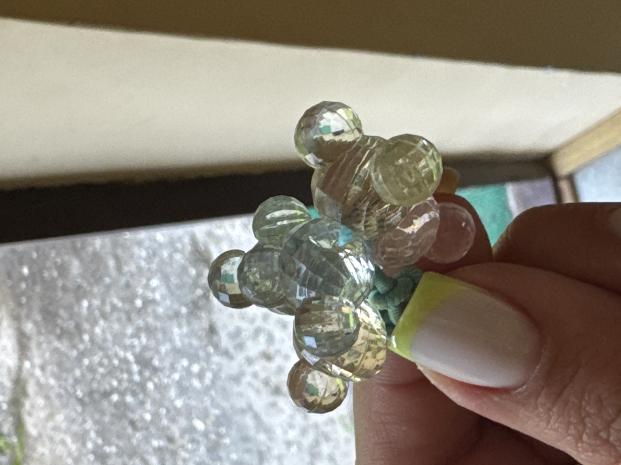 A close-up shot shows a hand holding a small, clear beaded decoration. The decoration is clustered and appears to be shaped like a flower or a small animal, with several faceted beads reflecting light. The hand has a manicured nail with a French tip and a bright yellow accent on the cuticle area. In the background, a blurred outdoor scene with a light-colored, textured surface and hints of greenery is visible. The right side of the image shows a plain, light-colored wall and a dark vertical shadow. The scene is well-lit, suggesting it was taken during the day, possibly outdoors or near a window. The location of Naro-Fominsk, Russia, is not directly depicted or inferable from the visual elements in the image.