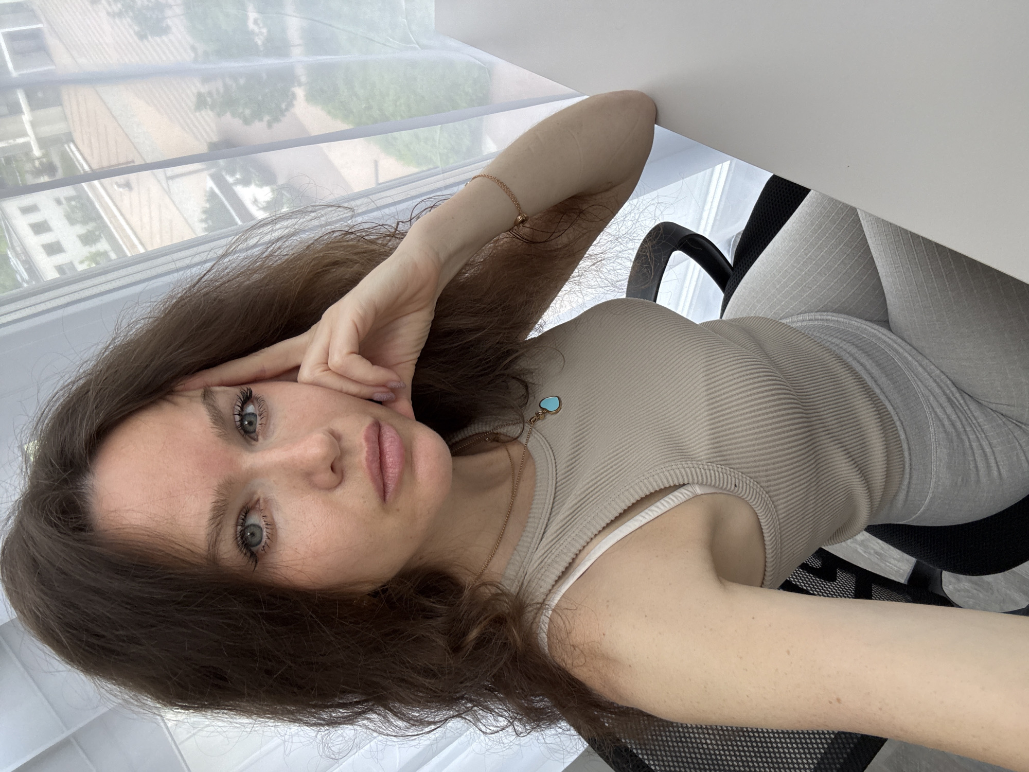 A young woman with long, wavy brown hair and blue-green eyes captures a selfie indoors, seated at a desk. She wears a light beige ribbed tank top over a white bra or camisole and light grey patterned leggings. Her right hand is positioned near her face, with fingers lightly touching her temple, while a delicate gold chain bracelet adorns her right wrist. A gold necklace with a small, heart-shaped turquoise pendant rests on her chest.
The woman is seated in a black mesh office chair, with a plain white desk surface visible in the foreground. Behind her, a large window reveals a bright, daytime urban or suburban scene, with blurred buildings and trees visible through sheer white curtains or blinds. The overall lighting is soft and bright, suggesting natural light. Her expression is calm and neutral, with a slightly pensive gaze directed towards the camera. No discernible text is visible within the image.