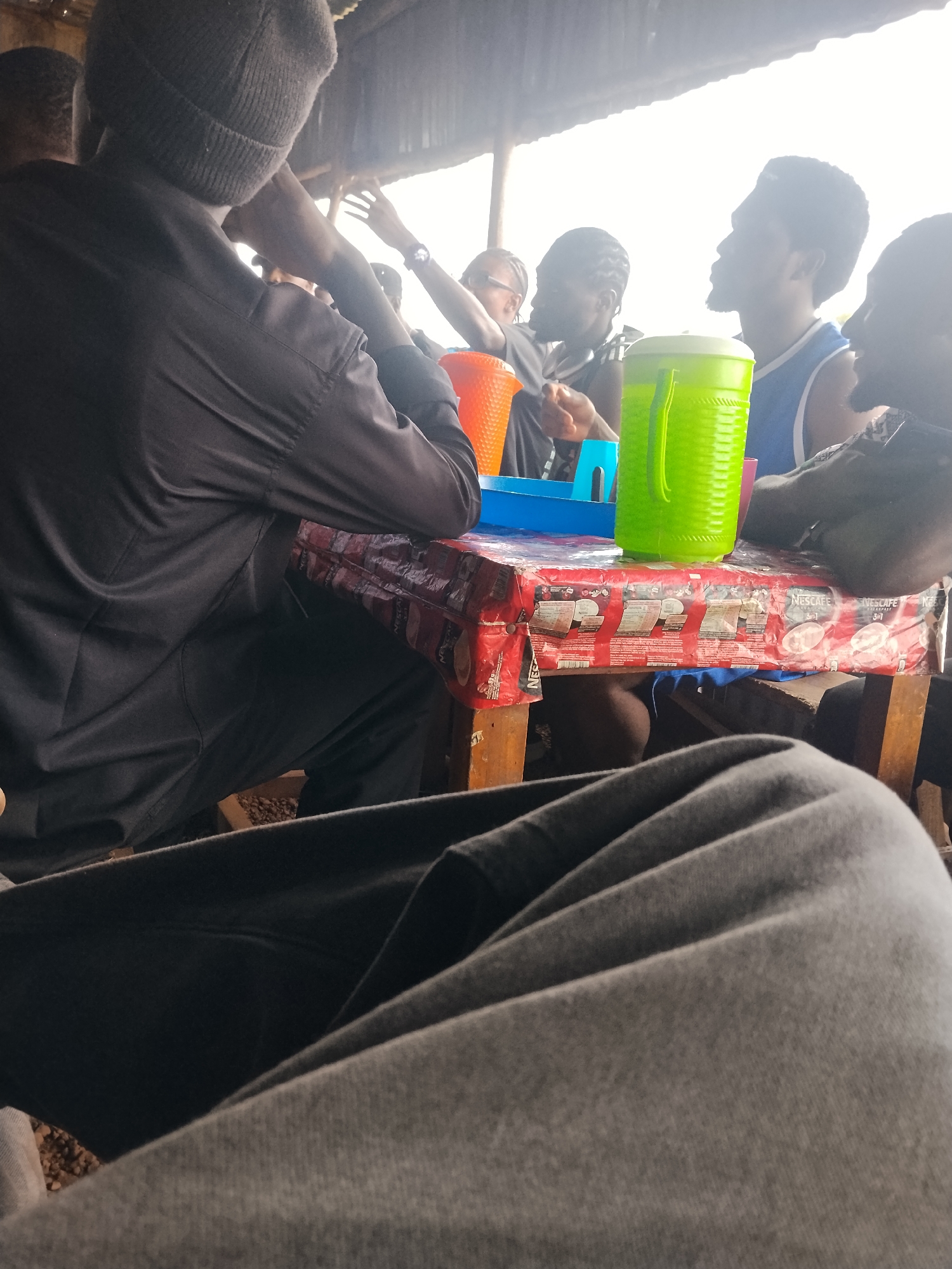The image depicts a group of individuals gathered around a table under a shelter, likely a roadside eatery or a casual meeting spot in Durumi, Nigeria. The foreground is dominated by the folded fabric of gray trousers, suggesting the perspective of someone seated. In the mid-ground, a wooden table is covered with a red and white Nescafé branded plastic sheeting. On the table, a bright green plastic pitcher and an orange plastic cup are visible. Several people are seated around the table, their faces mostly obscured by the angle and lighting. One individual, wearing a dark long-sleeved shirt and a grey beanie, is seen from the back. Another person is wearing a blue tank top. The background shows a corrugated metal roof and bright, overcast sky. The overall atmosphere appears casual and social, with people engaging in conversation or simply sharing space.