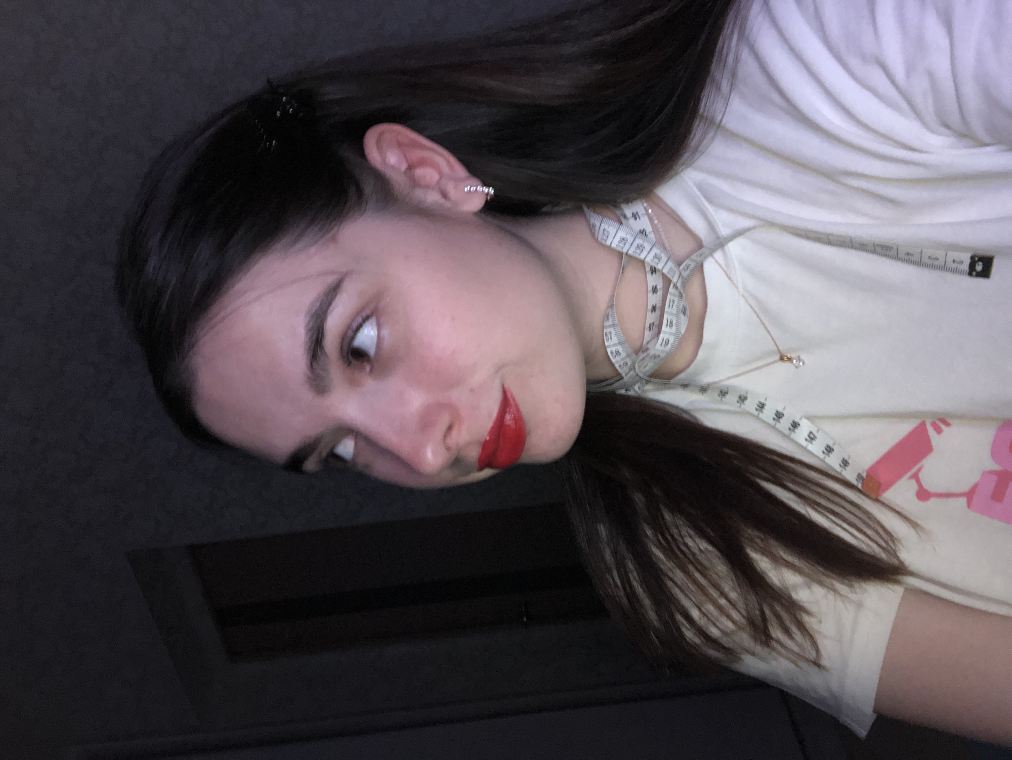 A young woman is seen at a sharp angle from below and to the side. She has dark hair, a fair complexion, and is wearing red lipstick. A measuring tape is draped around her neck, and she has a small silver stud in her ear. She is wearing a light-colored T-shirt. The background is a dark, indistinct surface. The location is not specified in the image and cannot be determined from the visual information provided.