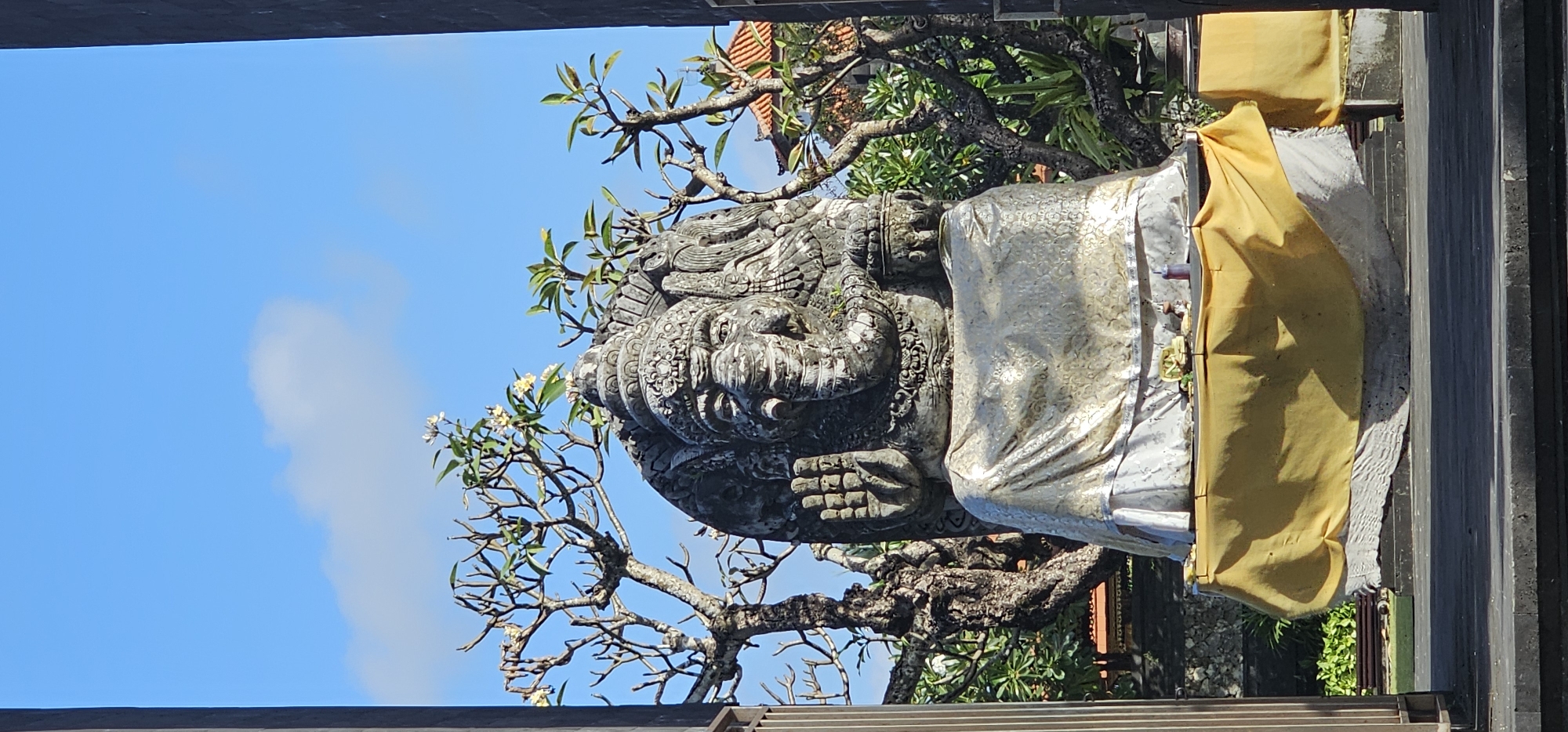 Captured from a low angle, the image portrays a Hindu statue in Denpasar, Indonesia. The statue is intricately carved from stone and is partially draped with white and yellow cloths. The statue is set amidst a backdrop of lush green foliage and a clear blue sky with some clouds. A portion of a wooden or stone structure frames the top and right side of the view, suggesting the statue is being viewed from a sheltered area.