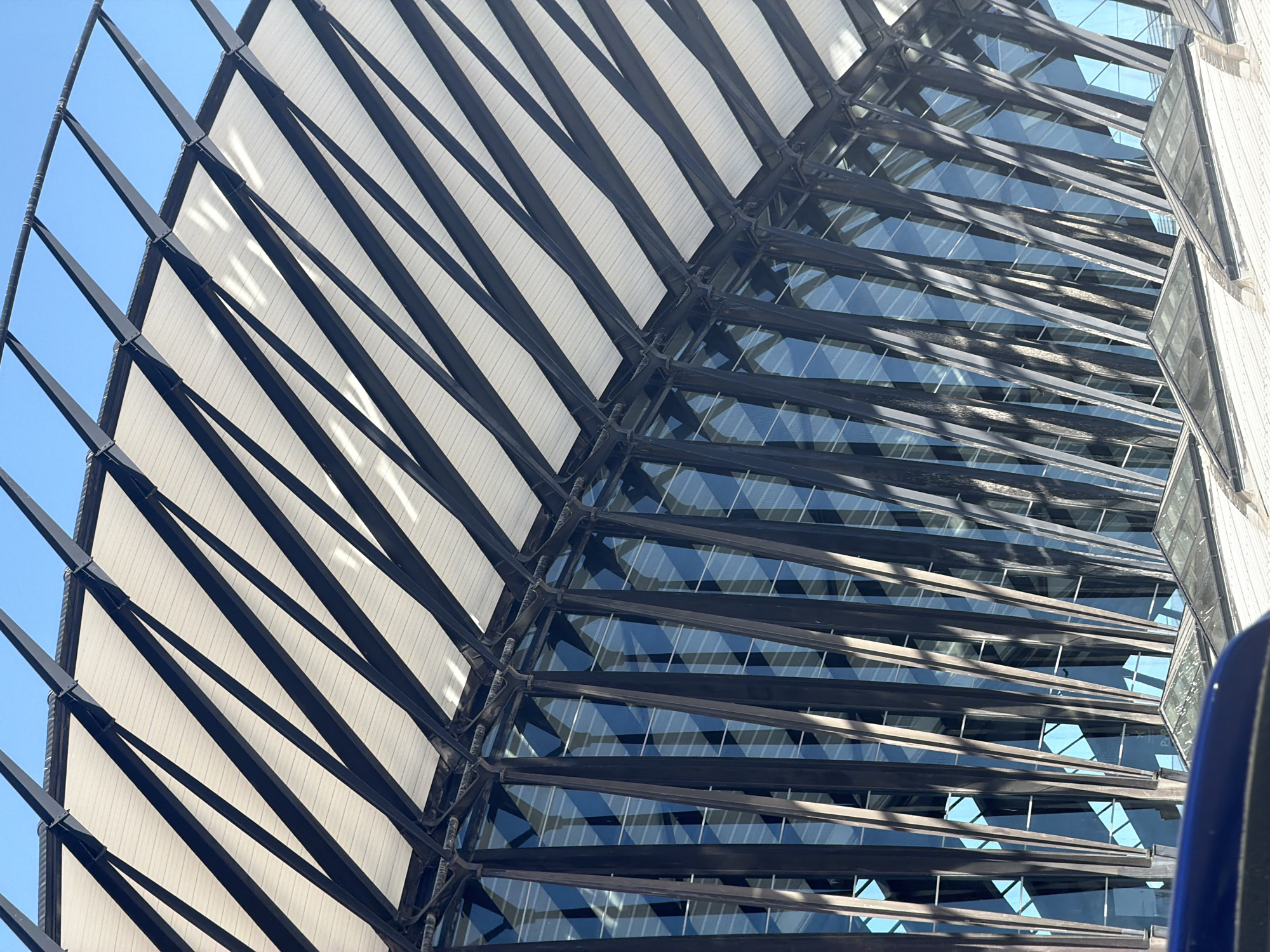 This is a low-angle, close-up, outdoor shot of the exterior of Lyon-Saint Exupéry Airport's Terminal 1 in Colombier-Saugnieu, France. The image focuses on the distinctive architectural elements of the building's roof and facade. The roof is characterized by a series of parallel, dark-colored, angled beams that support a white, textured surface, creating a ribbed effect. These beams cast shadows on the white material, adding depth and dimension.
Below the roof, the building's facade is composed of numerous vertical, dark-colored structural elements that are angled inwards and appear to be supporting the roof structure. These elements are interspersed with large glass panels that reflect the bright blue sky, creating a grid-like pattern of blue and dark vertical lines. The glass panels also appear to have some internal structural elements or reflections, adding to the complexity of the facade.
The foreground of the image shows a curved, dark blue object, possibly the hood of a vehicle, indicating the viewer is at ground level or within a vehicle looking up at the airport structure. Further down, and partially obscured by the blue object, are triangular glass skylights integrated into a white, textured rooftop or walkway area.
The overall impression is one of modern, large-scale architecture, emphasizing geometric patterns, strong lines, and the interplay of light and shadow. The scene is clean and orderly, suggesting a well-maintained public space. There are no people visible in the frame. The time of day appears to be daytime, given the bright sunlight and clear blue sky.