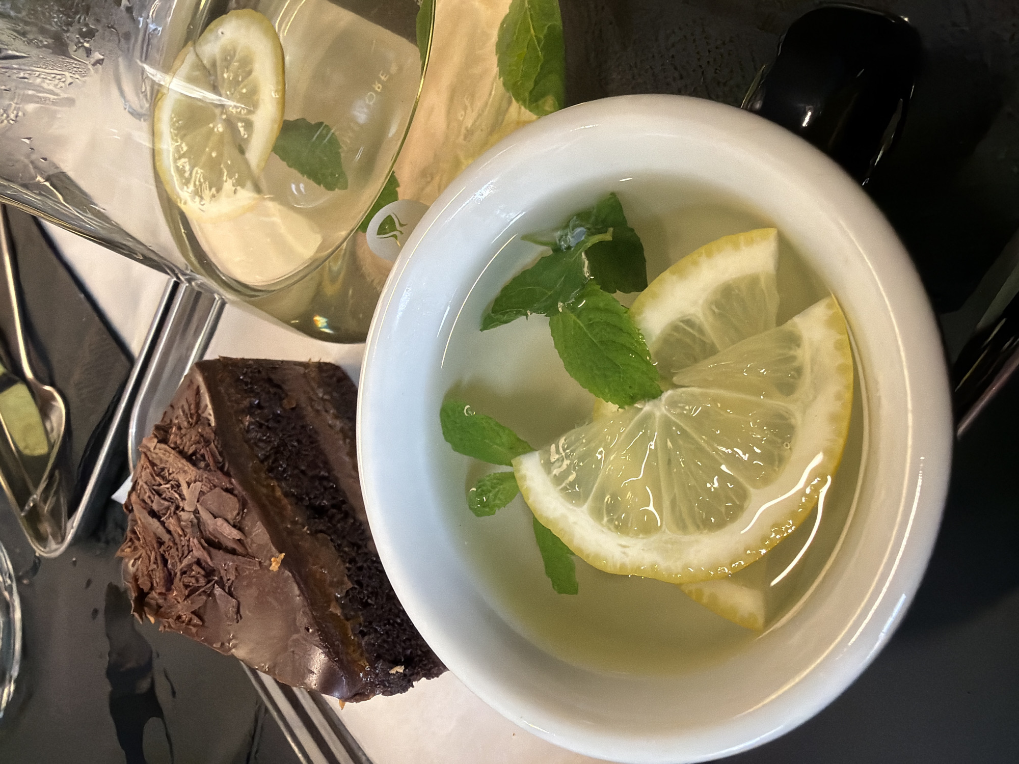 A high-angle shot depicts a slice of chocolate cake, a glass pitcher with lemon slices and mint, and a white cup also containing water, lemon slices, and mint leaves. The composition suggests a restaurant or cafe setting. The items are arranged on a black surface. No people are visible in the frame. The location is not possible to determine from the visual content.