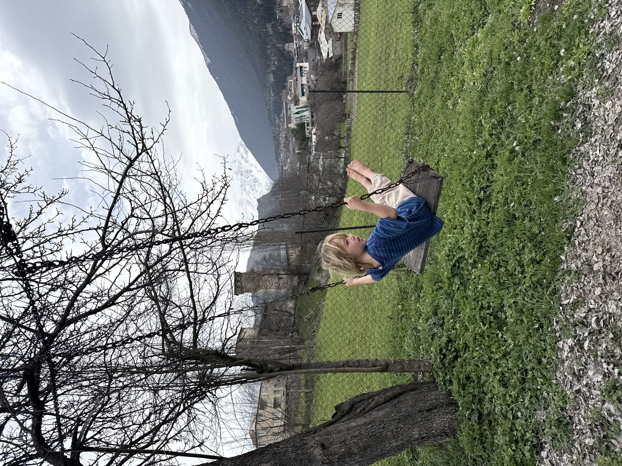 A child is on a swing in a grassy area. A bare tree is to the left. A chain link fence is visible behind the swing. In the background are buildings and a mountain covered with snow. The location is likely the city of Mestia, Georgia. The child is wearing a blue striped shirt and light-colored pants. The child is barefoot.