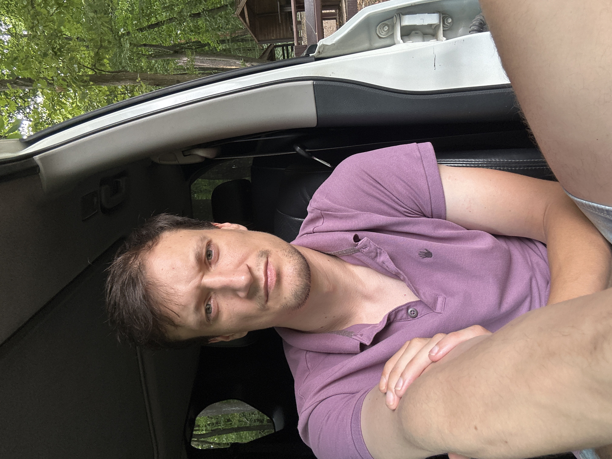 A man with short brown hair and a light beard is sitting in the driver's seat of a white vehicle. He is wearing a purple polo shirt and has his left arm resting on his right arm. His face is turned towards the camera, and he has a serious expression. The car appears to be parked outdoors, with trees and a small wooden structure visible in the background. The location is not Shatava, Ukraine, but rather a forested area.
