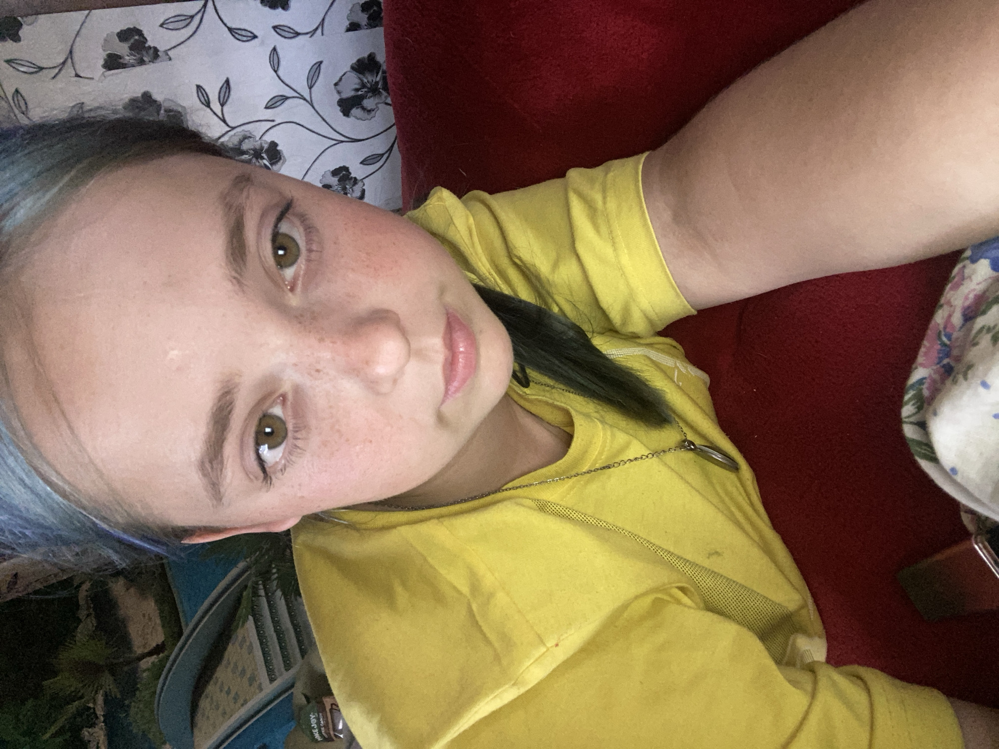 A young girl with blue-streaked hair is pictured in a close-up selfie. She is wearing a bright yellow t-shirt and a silver necklace. Her face is turned slightly towards the camera, and she has a neutral expression. Freckles are visible across her nose and cheeks. The background is blurred but appears to include a floral wallpaper and a red surface, possibly upholstery. To the left of the girl, a glimpse of an outdoor scene with palm trees and a blue structure, possibly a pool, can be seen. The location is not specified as Nova Praha, Ukraine.