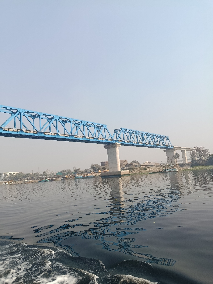 The image displays a wide, calm body of water, likely a river, under a clear, pale blue sky. Dominating the upper mid-ground is a bright blue metal truss bridge, extending horizontally across the frame. The bridge is supported by robust, light-colored concrete piers that rise from the water. The structure appears to be a multi-span bridge, with two distinct truss sections visible, and a further concrete segment extending towards the right on the far bank.
The foreground of the water shows ripples and white foamy wake, indicating that the photograph was taken from a moving boat or vessel. The water surface reflects the blue of the bridge and the light of the sky, creating shimmering patterns. Along the distant shoreline, behind the bridge piers, there's a low-lying landscape featuring various buildings, green trees, and some scattered vegetation. Several small boats or structures can be seen along the riverbank.
The time of day appears to be daytime, with bright, consistent lighting and no harsh shadows. The weather is clear, with a vast, unobstructed sky. There is no visible text in the image. This scene provides a tranquil view of a bridge and river landscape in Zazira, Bangladesh.