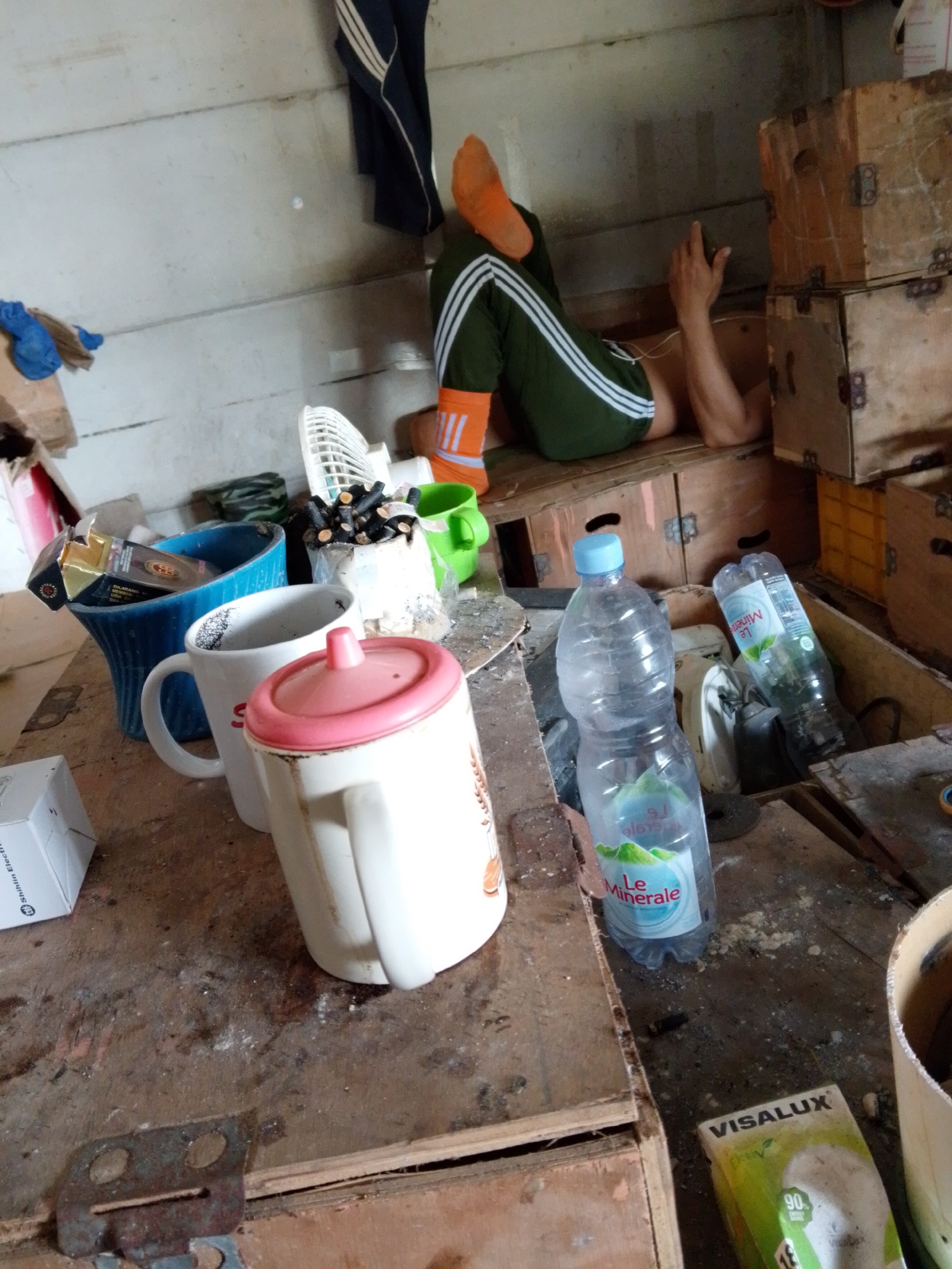 The scene appears to be an untidy indoor space in Bukit Ketok, Indonesia. The background shows a person wearing dark green athletic pants with three white stripes and orange socks, reclining against stacked wooden crates, possibly taking a rest. A dark blue tracksuit jacket hangs on the wall behind them. The foreground presents a cluttered surface covered in debris, including a white mug with a pink lid, a white mug, a blue vase holding a pack of cigarettes, a container full of cigarette butts, two plastic water bottles branded "Le Minerale," and a light bulb in a "VISALUX" package. Also visible are several wooden crates and other objects, creating a disorganized and unhygienic setting.