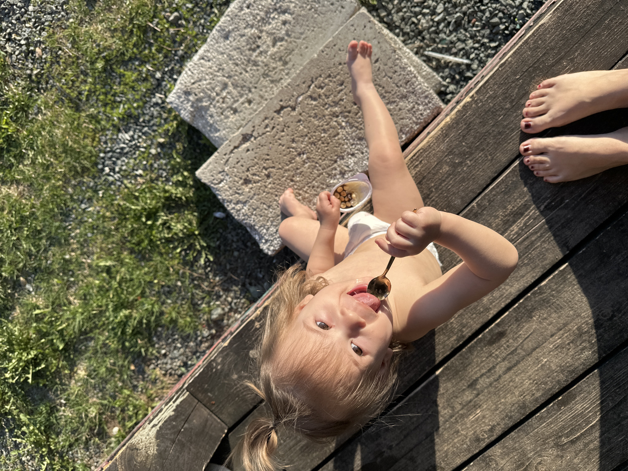 A high-angle outdoor photograph shows a young child sitting on a weathered wooden surface, possibly a deck or bench. The child, wearing light-colored bottoms, holds a spoon with food near their mouth, their tongue slightly extended as if about to eat. Beside the child is a small container filled with what appears to be pieces of food. The child has fair hair pulled into pigtails and is looking up towards the camera.
Next to the child, resting on concrete blocks, is a set of child's legs, which appear to be on the top of the blocks.
Adjacent to the wooden surface and concrete blocks, the ground is covered in grass and gravel.
On the opposite side of the child are the feet of an adult, positioned on the wooden surface with toes painted red.
No other information suggesting a specific location, such as Kharnavy, Russia, is evident in the image. The scene appears to be in a residential or park setting.