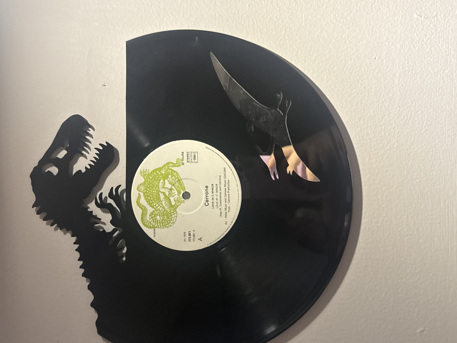 A vinyl record is affixed to a white wall, featuring a black dinosaur silhouette on its left. The record label displays "Cerrone" and "LOVE IN C MINOR," indicating the artist and song title, along with 1976 copyright information. A cut-out of a black Pterodactyl appears on the right side of the record, with an unusual iridescent reflection on its bottom half.