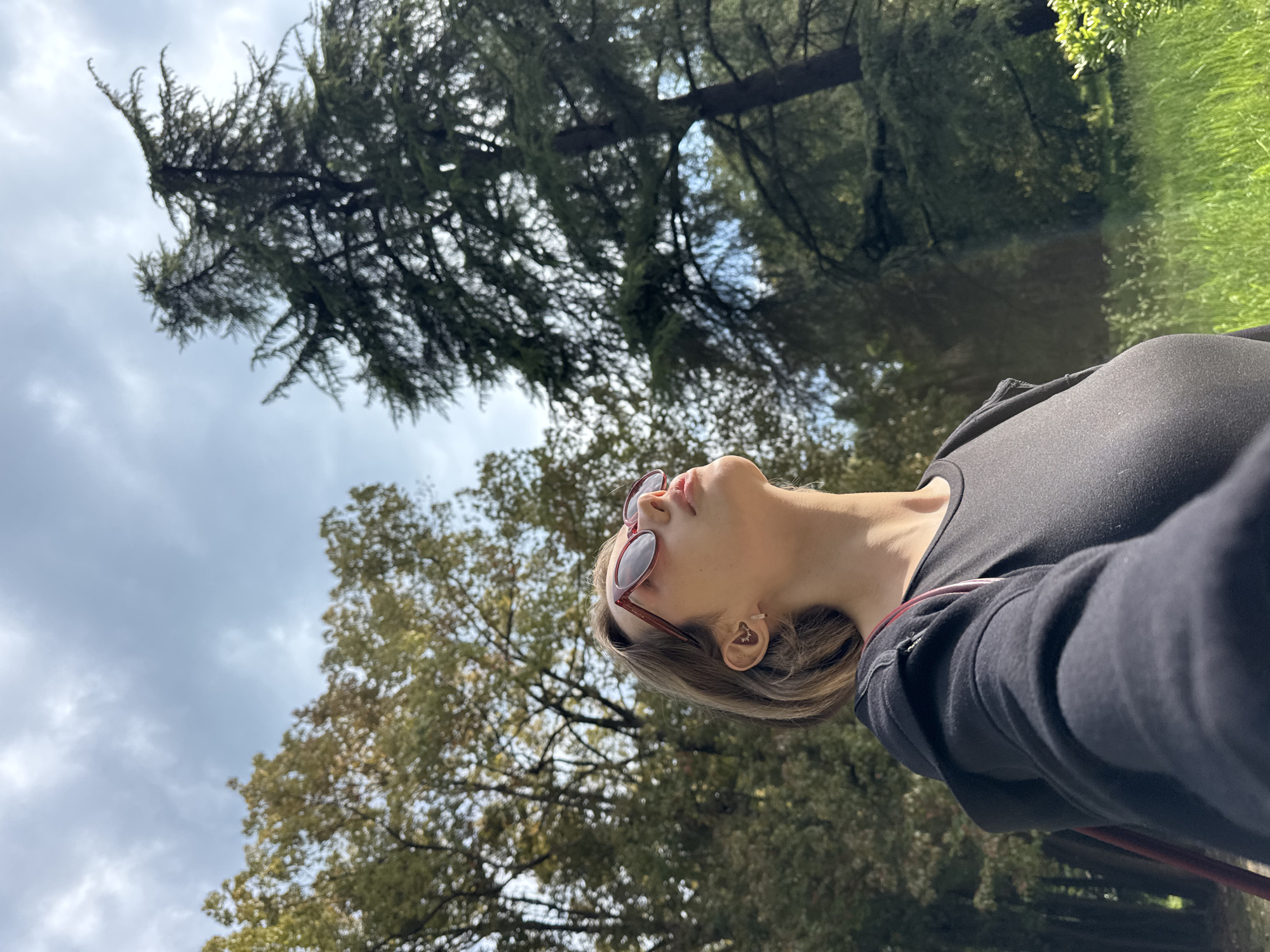 The photograph presents a low-angle view of a person outdoors, their head tilted upwards. The individual is wearing sunglasses with a red frame and a dark-colored top, possibly a hoodie. Their fair skin and light brown hair are visible. The backdrop consists of a partly cloudy sky and trees with dense foliage. The photograph appears to be taken from a position looking upwards at the subject and the surrounding trees. The location is indeterminate.