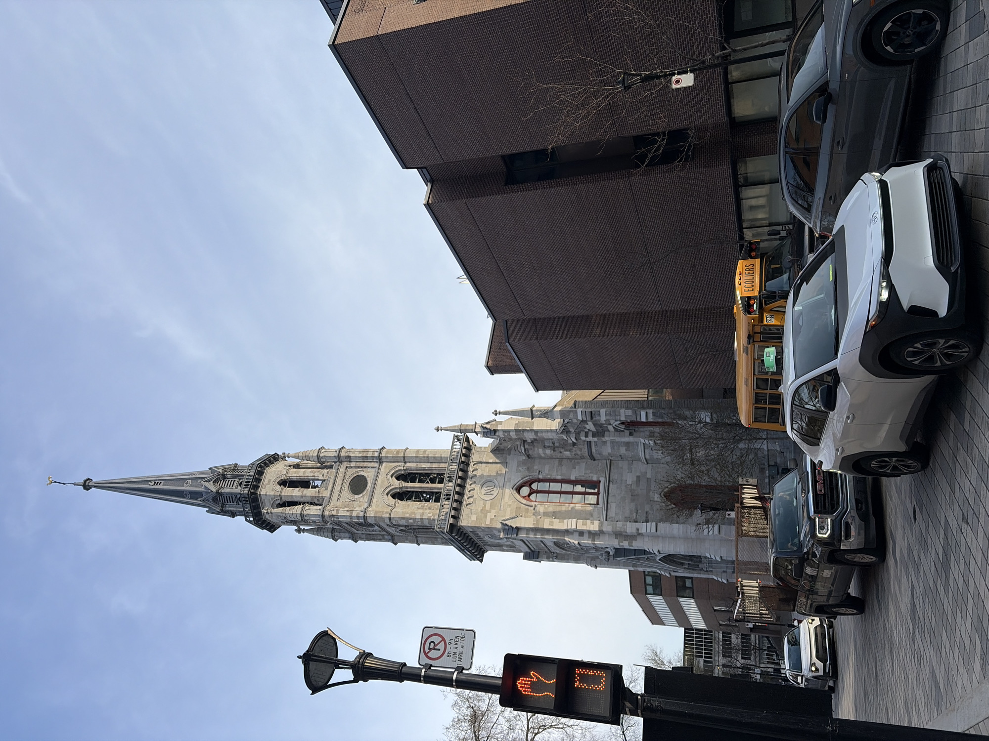 The image captures a street scene in Montreal, Canada, featuring the iconic spire of Notre-Dame Basilica. The camera angle is low, looking upwards towards the spire, which dominates the left side of the frame against a pale blue sky. The church structure is made of grey stone, displaying detailed architectural features, including arched windows and intricate stonework.
On the right side of the image, modern brick buildings and a line of parked vehicles, including a white SUV, a dark pickup truck, and a yellow school bus, line the street. A traffic light displaying a "Don't Walk" signal and a "No Parking" sign are visible on a street post in the foreground. The sidewalk is paved with patterned bricks. The scene appears to be captured during daylight hours.