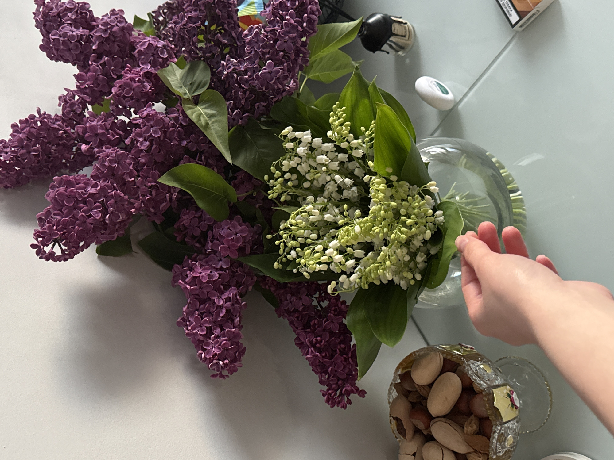 An overhead shot captures a vibrant bouquet of flowers, featuring dense clusters of deep purple lilacs alongside delicate white lily of the valley blossoms, arranged in a clear glass vase on a light, reflective surface. A fair-skinned hand reaches into the frame from the right, positioned near the vase.
To the lower right, a small, ornate clear glass bowl, possibly crystal, is filled with nuts, predominantly shelled and unshelled pistachios. In the background, scattered on the surface, are several objects: a small black and white object resembling a mini football or rugby ball, a smooth white oval item, and a rectangular package, possibly a cigarette pack, partially visible with faint text "26484 A" on it. The reflective surface creates subtle reflections of the flowers and other items. The lighting suggests an indoor setting during the day.