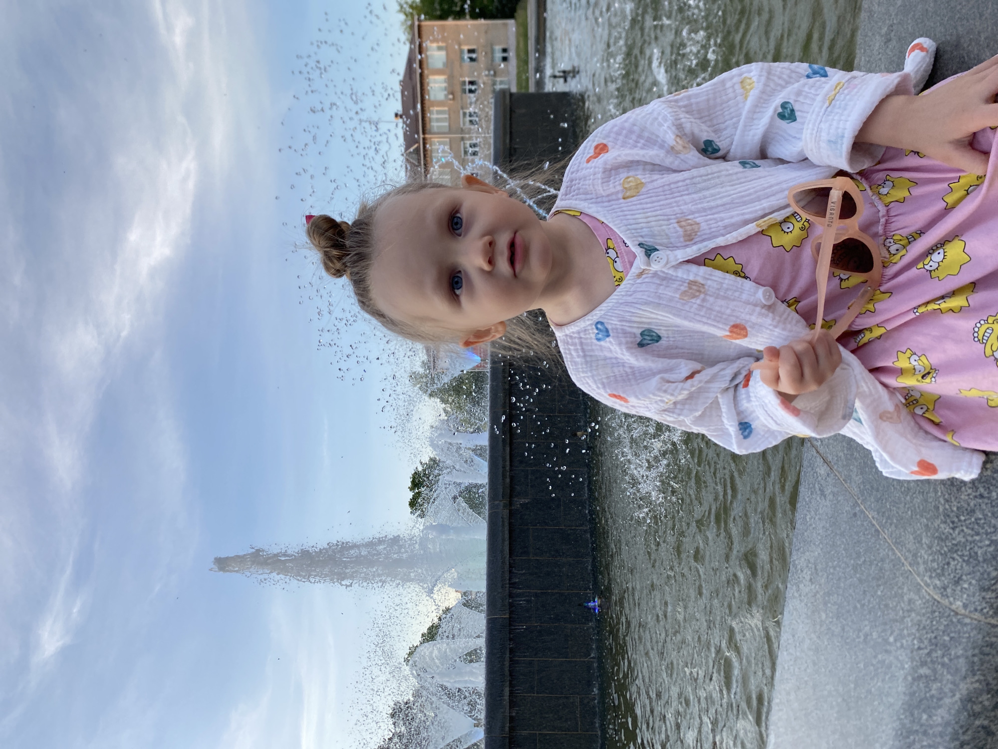 A young girl with blonde hair pulled up in a bun stands in front of a fountain in Chaikovsky, Russia. She is wearing a pink dress with a Lisa Simpson pattern and a white cardigan with colorful heart and star designs. She holds pink sunglasses in her left hand. The fountain is black and the water is shooting upwards in multiple streams, some of which are spraying towards the girl. In the background, a building is visible. The sky is light blue with some white clouds.