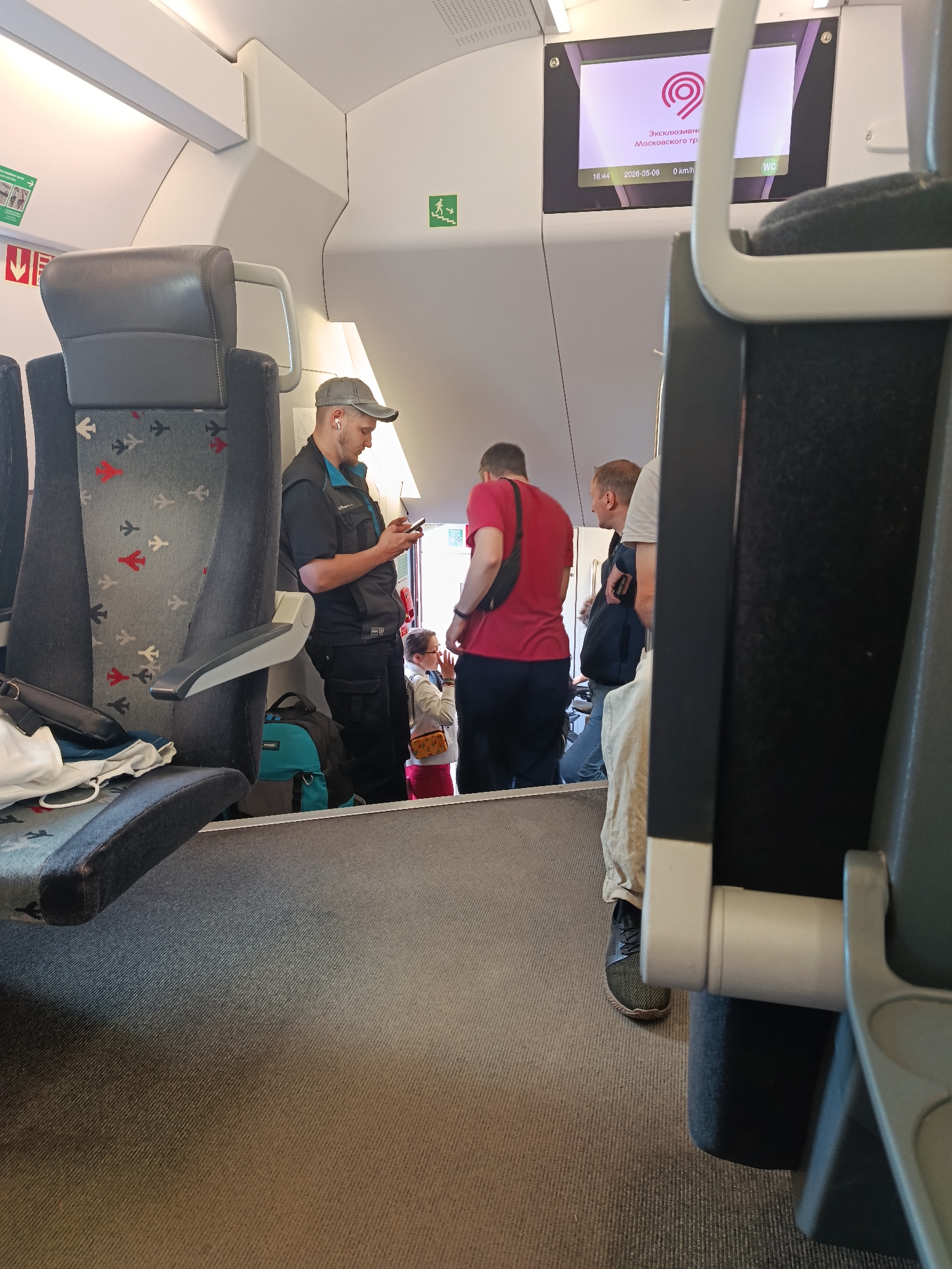 The image was taken inside a train car, showing several people near an open doorway. One man in a gray hat and dark vest is using a mobile phone, while another man wearing a red shirt stands nearby. A train seat with an airplane pattern is visible in the foreground. A screen on the wall displays the text "Эксклюзивно Московского тр".