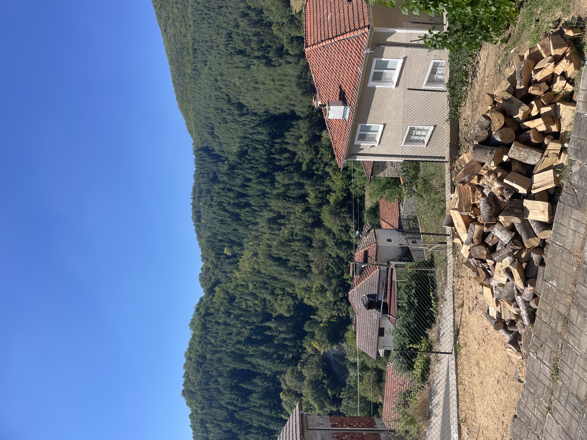 This is a sunny, vertical, medium-shot photograph of a rural village nestled at the base of a densely forested mountain range. The sky is a clear, vibrant blue, occupying the top third of the image. The foreground is dominated by a large pile of cut firewood, composed of various sizes and types of logs. The firewood is situated on a patch of sandy earth, with a paved area and some concrete steps visible in the bottom left corner. Behind the firewood, a chain-link fence delineates a property line.
Beyond the fence, several houses with red-tiled roofs are visible. One house, in the mid-right of the frame, is light-colored with white-framed windows and a chimney. Another house, partially visible on the far left, appears to be constructed of brick. Green foliage and some smaller trees are scattered amongst the houses.
The middle ground and background are dominated by a steep mountain covered in a thick, evergreen forest. The trees create a textured green carpet stretching upwards towards the sky. A few patches of lighter colored earth or rock are visible on the mountainside, breaking the uniformity of the forest. The overall impression is one of a peaceful, natural setting in a mountainous region. There are no people or animals visible in the image. The location is identified as Oryahovets, Bulgaria, suggesting a setting within this region.