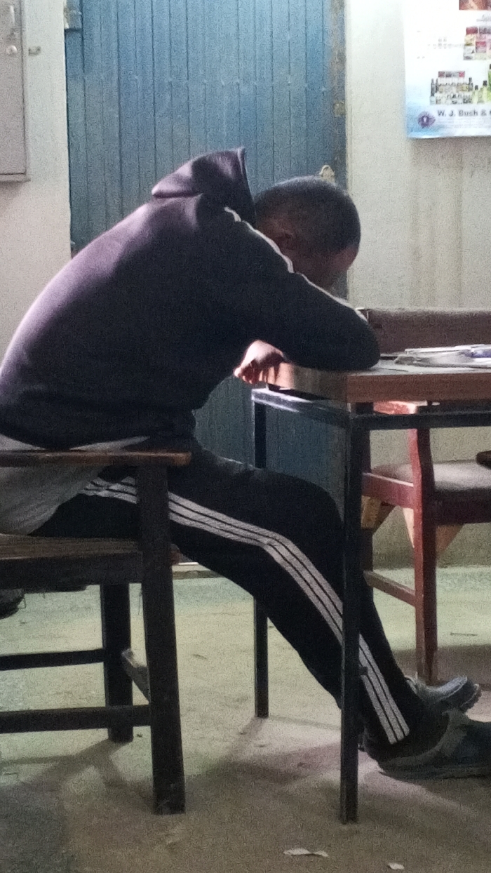 An indoor scene, possibly within Dakata, Nigeria, features a single individual seated at a table. The person, appearing to be an adult male, wears a dark hooded sweatshirt and dark track pants with three white stripes along the outer leg seams, coupled with dark footwear. He is positioned on a wooden chair with dark metal legs, leaning forward with his head resting on his forearms, which are placed upon a light-colored wooden table with dark metal legs. Papers or documents are visible on the table surface to the right of the individual's arms. A second wooden chair with reddish-brown components is situated to the right of the table. The background includes a solid blue door directly behind the seated person and a light-colored wall to the right, displaying a poster with text, including "W. J. Busch," and various images. The floor surface is light-colored, indicative of concrete or tile.