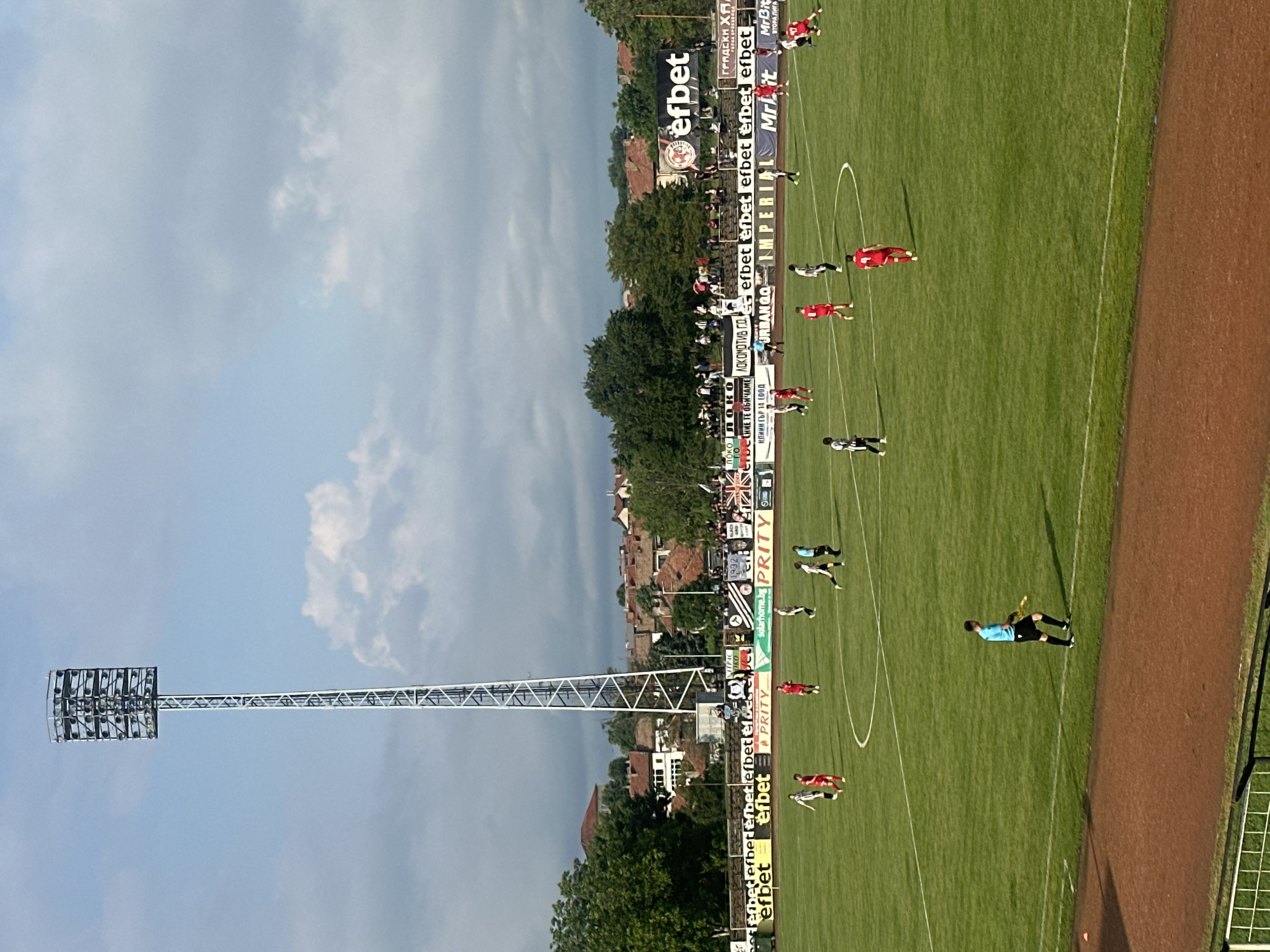 The image captures a soccer game in Gorna Oryahovitsa, Bulgaria. The view is from an elevated position, overlooking the field with players in red and white/black striped uniforms. A referee in a light blue shirt and black shorts is also visible. Advertising banners line the field, featuring brands like "efbet" and "PRITY," among others. The stadium has a low-rise brick building near the field perimeter. The sky is partly cloudy, and a tall floodlight structure is visible in the left part of the image. Spectators are present in the background.