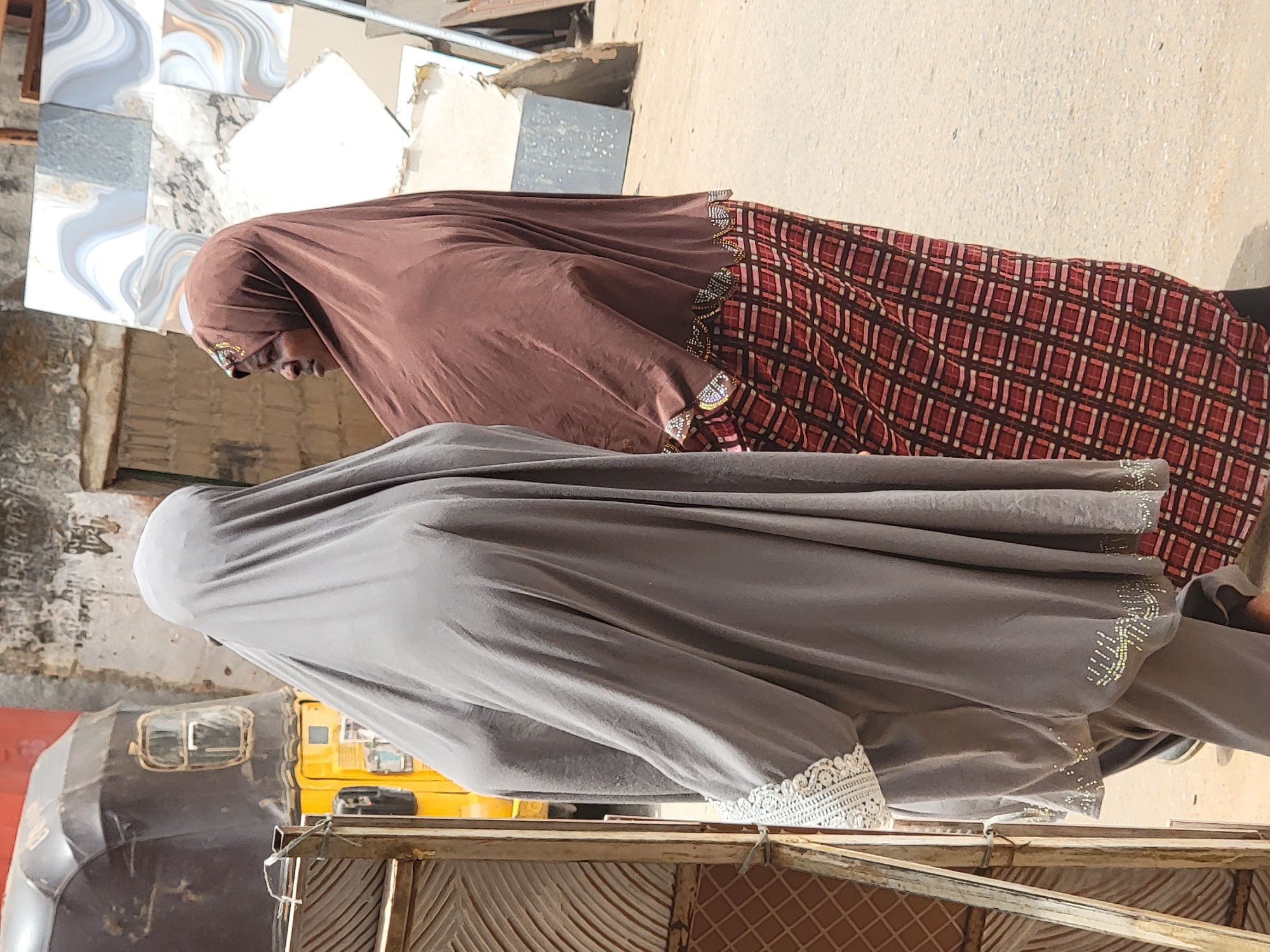 Two women wearing hijab are walking in Maiburiji, Nigeria. The woman in front is wearing a brown hijab with red and brown patterned fabric. The woman behind is wearing a gray hijab with white lace at the edge. In the background are construction materials, a yellow and black vehicle, and the side of a building.