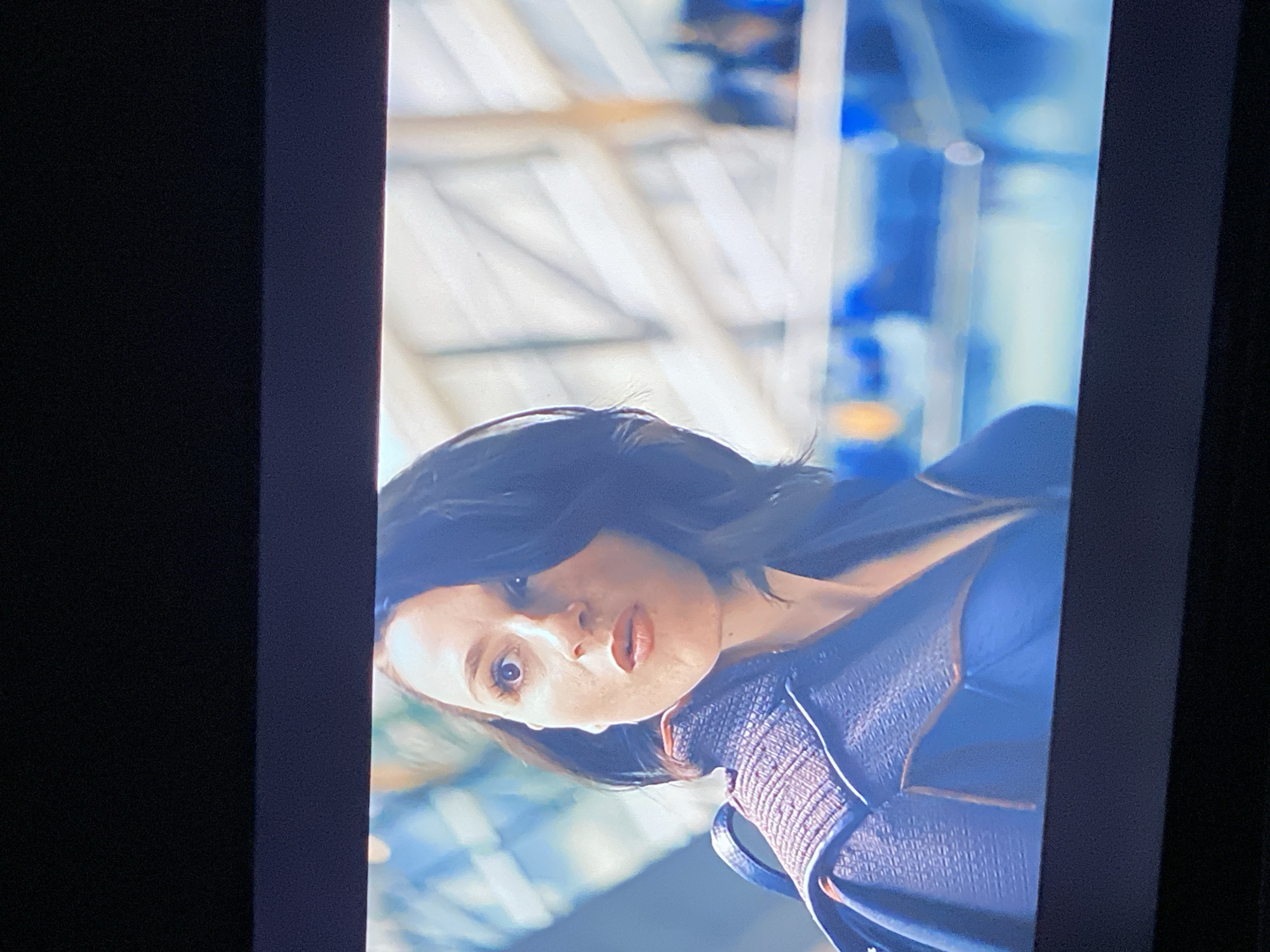 The image is a close-up shot of a woman with dark hair and fair skin. She is wearing a blue garment with a textured collar. Her expression is one of surprise or concern, with wide eyes and slightly parted lips. The background is blurred, suggesting a busy indoor environment with architectural elements like beams and railings, possibly an airport or a large public building. There are no clear indications of the city of Zastavna, Ukraine.