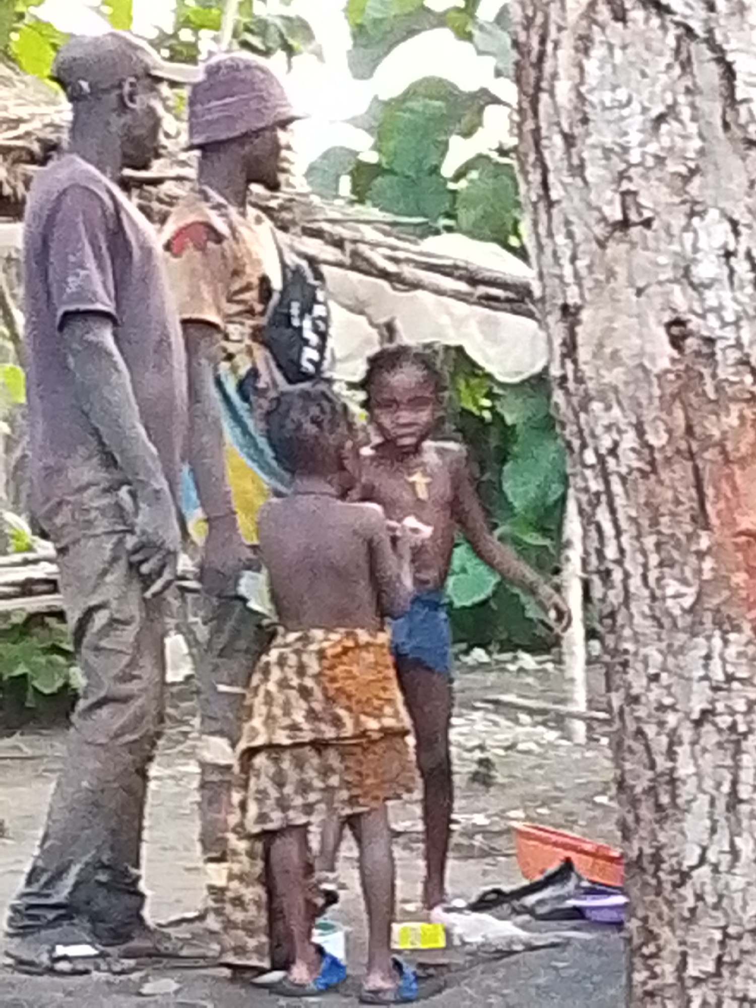 The image shows a group of people in an outdoor setting that appears to be somewhat rustic. In the foreground, there are two adults and two children. One adult on the left is wearing a purple t-shirt and pants, and a cap. Next to him, another adult is wearing a tan shirt and a bucket hat, with a bag slung over his shoulder. To the right of the adults are two children. The child closer to the foreground is wearing a leopard-print skirt and is shirtless. The child behind them is wearing blue shorts and is also shirtless. The background shows vegetation and some man-made structures that are indistinct. There is a large tree trunk on the right side of the image. The ground appears to be dirt or sand. The lighting suggests it is daytime. There are no obvious signs of urban infrastructure or specific landmarks that would indicate a city.