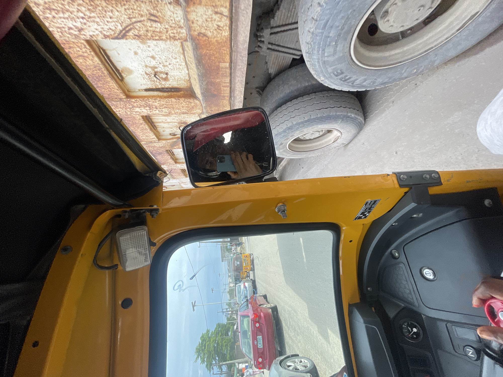 The image, taken from inside a yellow tricycle in Ado, Nigeria, is oriented with the right side facing up. Overhead are the rusty metal underside and the tires of a large vehicle. A side mirror reflects the photographer and their phone. Through the tricycle's window, a street scene is visible. Cars and other yellow tricycles are on the road. The sky is partly cloudy. The tricycle's dashboard and steering controls are on the right side of the image, with a person's hand visible near the steering wheel.