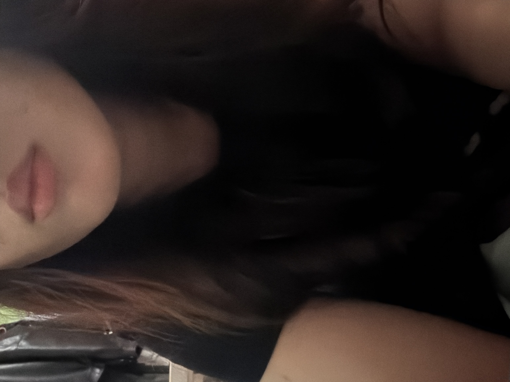 The image is a close-up, dimly lit shot of a person's face, focusing on their mouth and chin area. The person has light skin and dark hair that falls around their face. They are wearing a black t-shirt. The background is dark and out of focus, with some indistinct shapes that might be clothing or furniture. The image appears to be taken indoors, possibly in a bedroom or living space. There is no information available to determine the location in the city of Unknown, Russia.