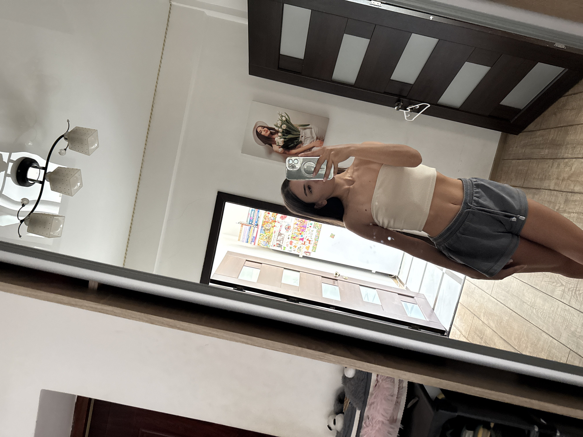 A young woman with long brown hair is taking a mirror selfie in a room. She is wearing a cream-colored strapless top and grey shorts. Her phone is held up to take the picture. In the background, a large mirror reflects part of the room, including a door with glass panels and a framed picture on the wall of a woman holding tulips. The floor is made of light-colored wood planks. There is a ceiling light fixture with multiple square shades. The overall impression is of a casual, indoor setting. The image does not provide any specific visual cues to determine its exact location in Chișinău, Moldova.