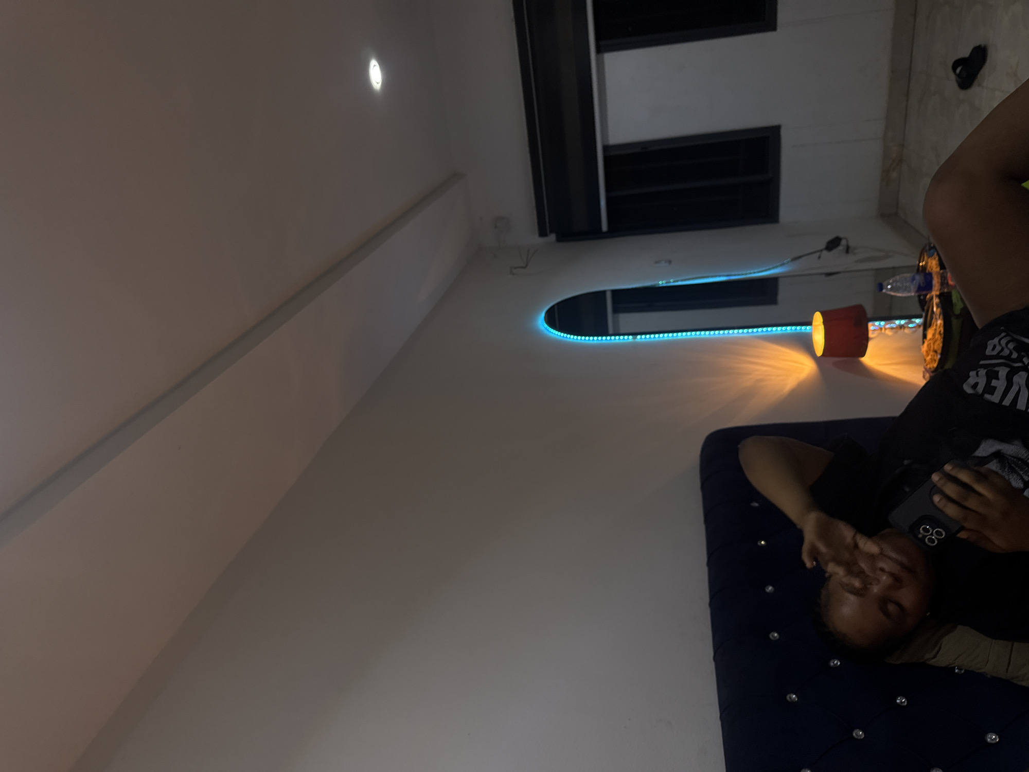 A person is lying on a dark blue tufted bed, holding a cell phone in their hand. The room has a window with horizontal blinds and a curved alcove with blue LED lighting. There's a small, cylindrical orange light on a nearby surface. A water bottle and a pair of black shoes are also visible in the background. The location is possibly in a residential setting in Abuja, Nigeria.