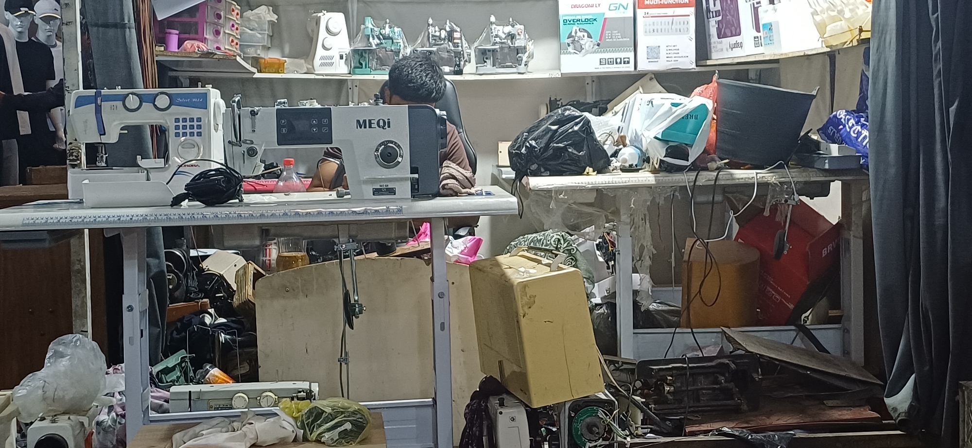The image depicts a cluttered workshop or store, likely in Булокчинор, Tajikistan, filled with sewing machines and related equipment. In the foreground, a white and blue sewing machine is positioned on a white table with a ruler marking. To its right, a silver and dark gray MEQI brand sewing machine is also on a table. A person, partially visible from the back and side, is seated at the MEQI machine, appearing to be operating it. The background reveals shelves stacked with various items, including boxes labeled with sewing machine brands like "DRAGONFLY" and "GN," as well as more sewing machines, some appearing older or disassembled. A large black trash bag and other plastic-wrapped items are piled on a table to the right of the MEQI machine. Other elements in the scene include a blue bucket, red boxes, and miscellaneous tools and materials scattered throughout the space. The overall impression is one of a working environment that is heavily utilized and somewhat disorganized.