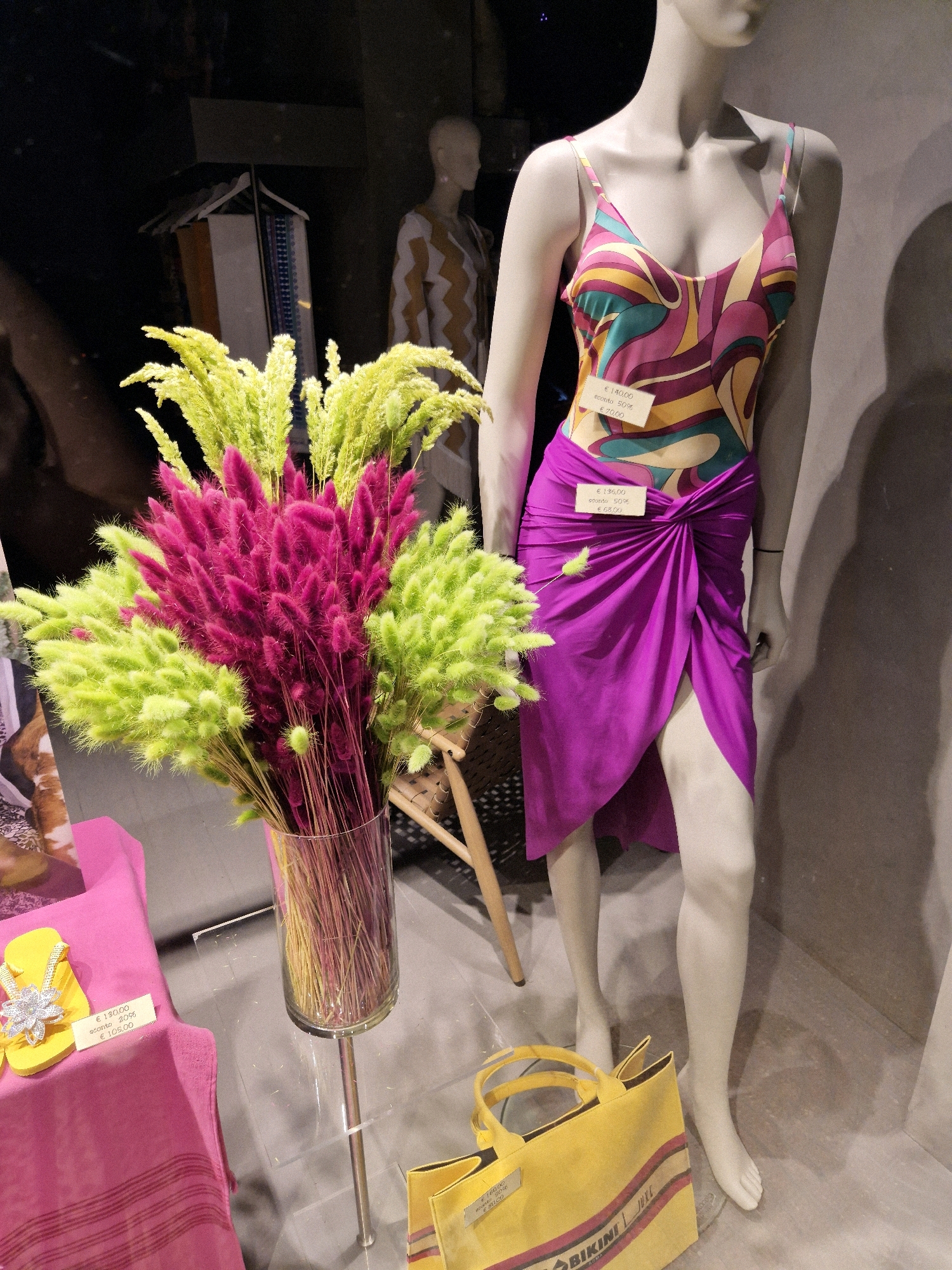 The image shows a store window display featuring a mannequin dressed in a colorful swimsuit and a purple sarong. A bouquet of dried flowers, with pink and green plumes, is arranged in a glass vase in front of the mannequin. To the left, a yellow flip-flop with a jeweled embellishment sits on a pink fabric. A yellow tote bag is visible on the floor in front of the mannequin. Several price tags with Italian text are attached to the clothing and the flip-flop. The background is dark and indistinct. The image is likely from a shop in Taranto, Italy, as indicated by the presence of Italian text on the price tags and the shopping bag.