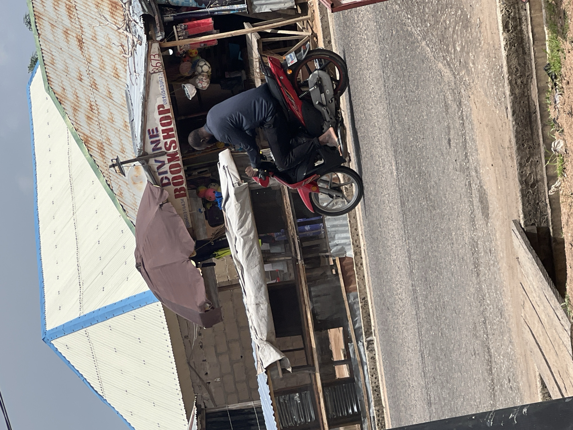 In Jalingo, Nigeria, a man rides a red motorcycle on a paved road. The man, with dark skin and a shaved head, is wearing a dark blue long-sleeved shirt and black pants. His left foot is bare. The motorcycle has a silver exhaust and red accents on the wheels. Behind him, a roadside stall features a sign that reads "DIVINE BOOKSHOP" in large red letters, with a phone number "08165438673" and the number "3673" displayed. Various items, including what appear to be footballs or decorative spheres, hang from the stall. A brown umbrella is positioned over the stall. The background shows buildings with corrugated metal roofs, one predominantly white and blue, and another with a rusty, weathered appearance. The environment appears to be a bustling urban or semi-urban area, with evidence of commercial activity and typical street infrastructure.