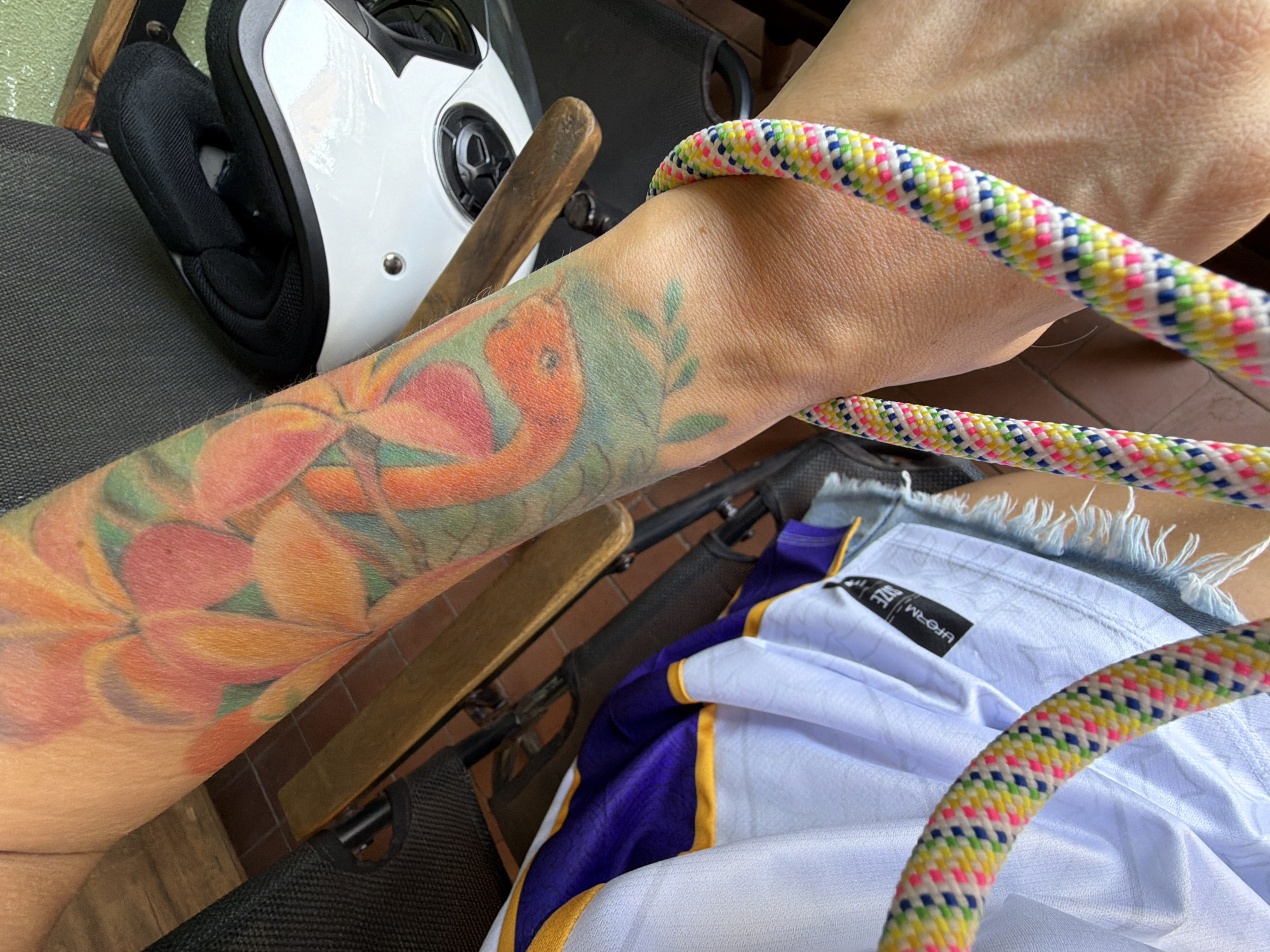 The image shows a person's arm with a colorful tattoo of flowers and a snake. The arm is resting on a wooden surface, possibly a table or chair. A white motorcycle helmet with black accents is visible in the background, along with a white and purple sports jersey. A brightly colored rope, possibly a climbing rope, is looped around the person's wrist and extends out of the frame. The scene appears to be outdoors, with a hint of a brick floor visible. The image does not provide any specific information about the location in Nha Trang Ward, Vietnam.