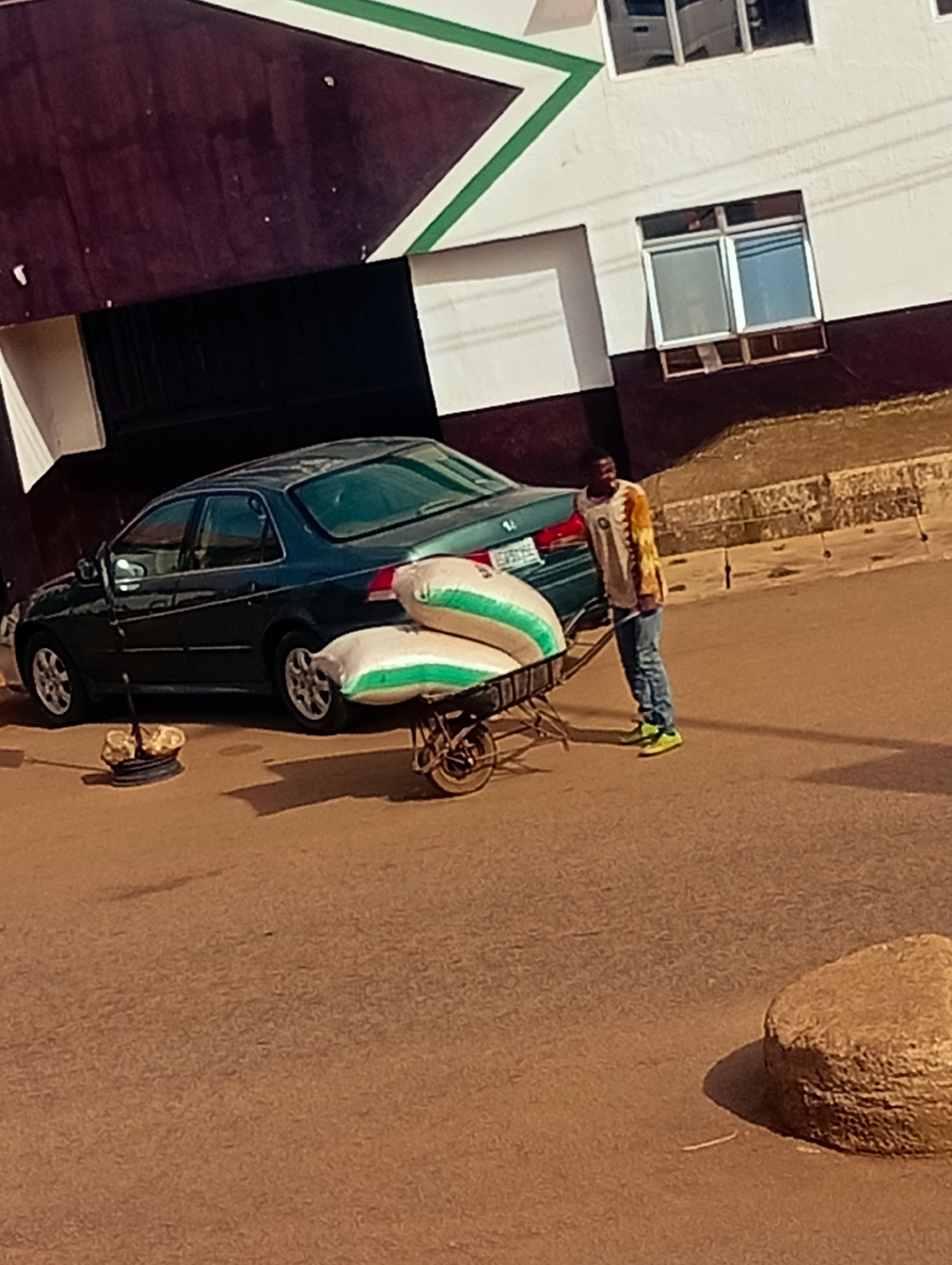 In Bukuru, Nigeria, a young man stands on a paved street next to a dark green sedan. He is wearing a long-sleeved, multi-colored shirt and blue jeans, with bright green sneakers. Beside him, a two-wheeled cart is loaded with two large white sacks, each with green stripes. The cart is positioned near the rear passenger side of the car. In the foreground, a rough, circular stone sits on the pavement. The background features a building with dark brown and white walls, adorned with green geometric accents. The sky is not visible.