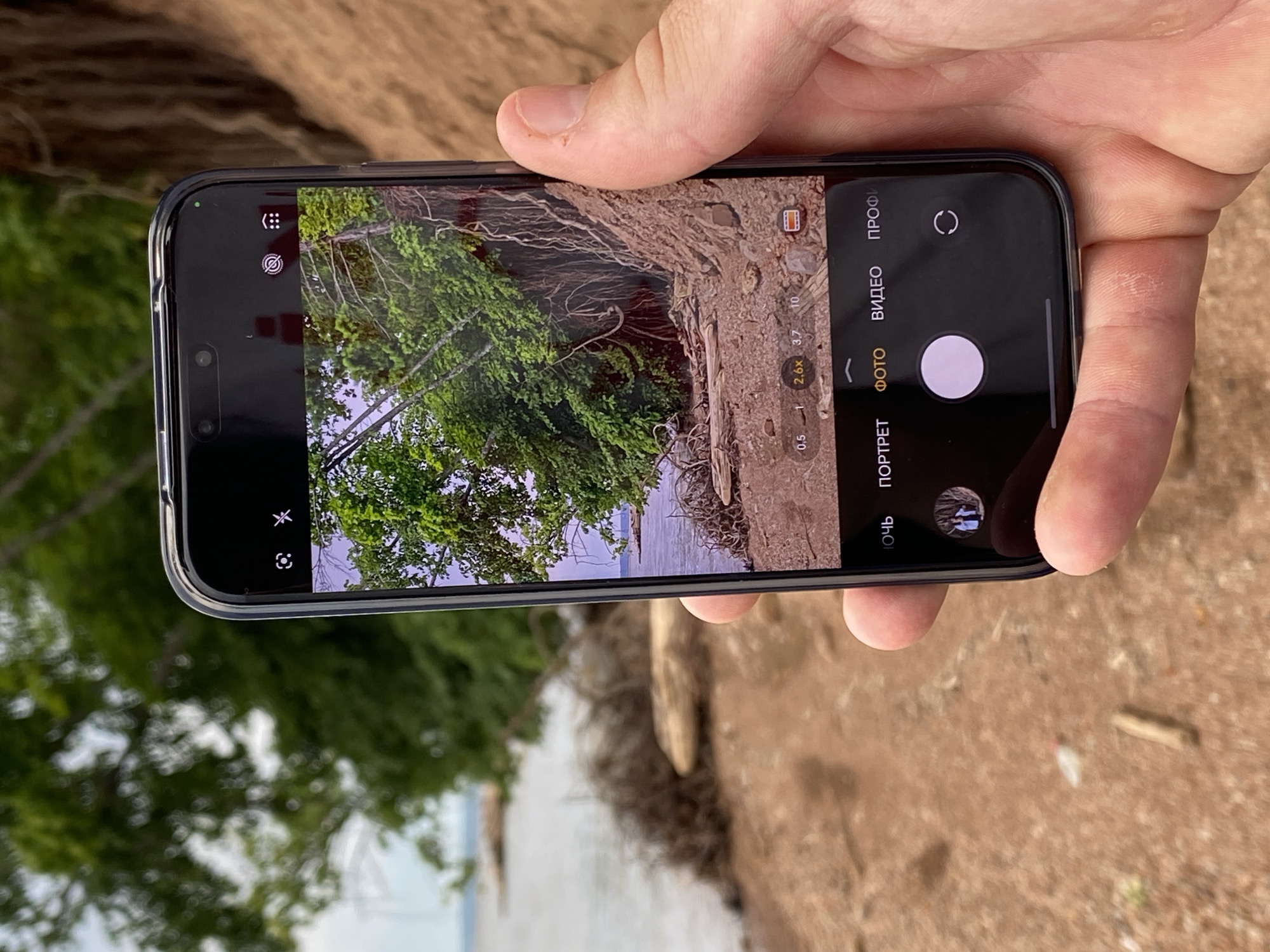 A person's right hand is holding a smartphone, capturing a view of the Kama River near the city of Chaikovsky, Russia. The phone screen displays the camera interface, with options for "NIGHT", "PORTRAIT", "PHOTO", "VIDEO", and "PRO". A zoom level of "2.6x" is indicated. The background shows the muddy bank of the river with green trees and vegetation, with a body of water visible in the distance. The sky is overcast.