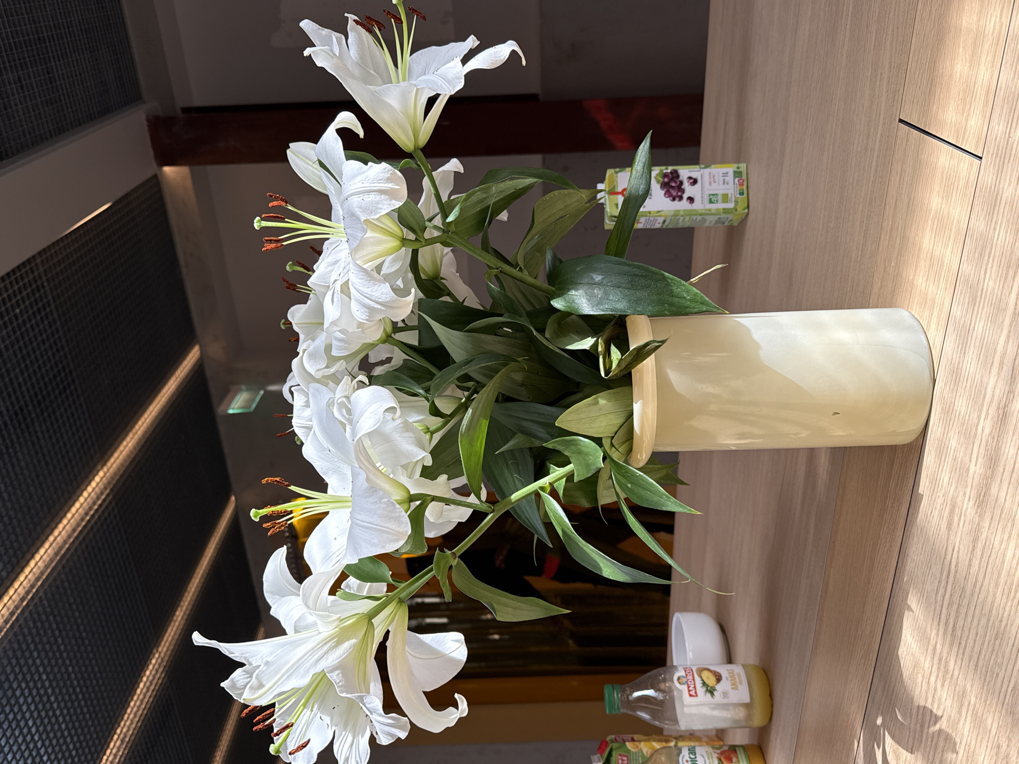 A still-life image depicts a bouquet of white lilies in a tall, cylindrical vase, placed on a light-colored wooden surface. The lilies are fully open, displaying their prominent stamens and pistils. The flowers are surrounded by green leaves. Near the vase's base are a small white cup and several bottles of juice.
The background includes a grey lattice wall, with a hint of exposed ceiling beams. The juice is from the brand Andros. The exact location is not indicated.