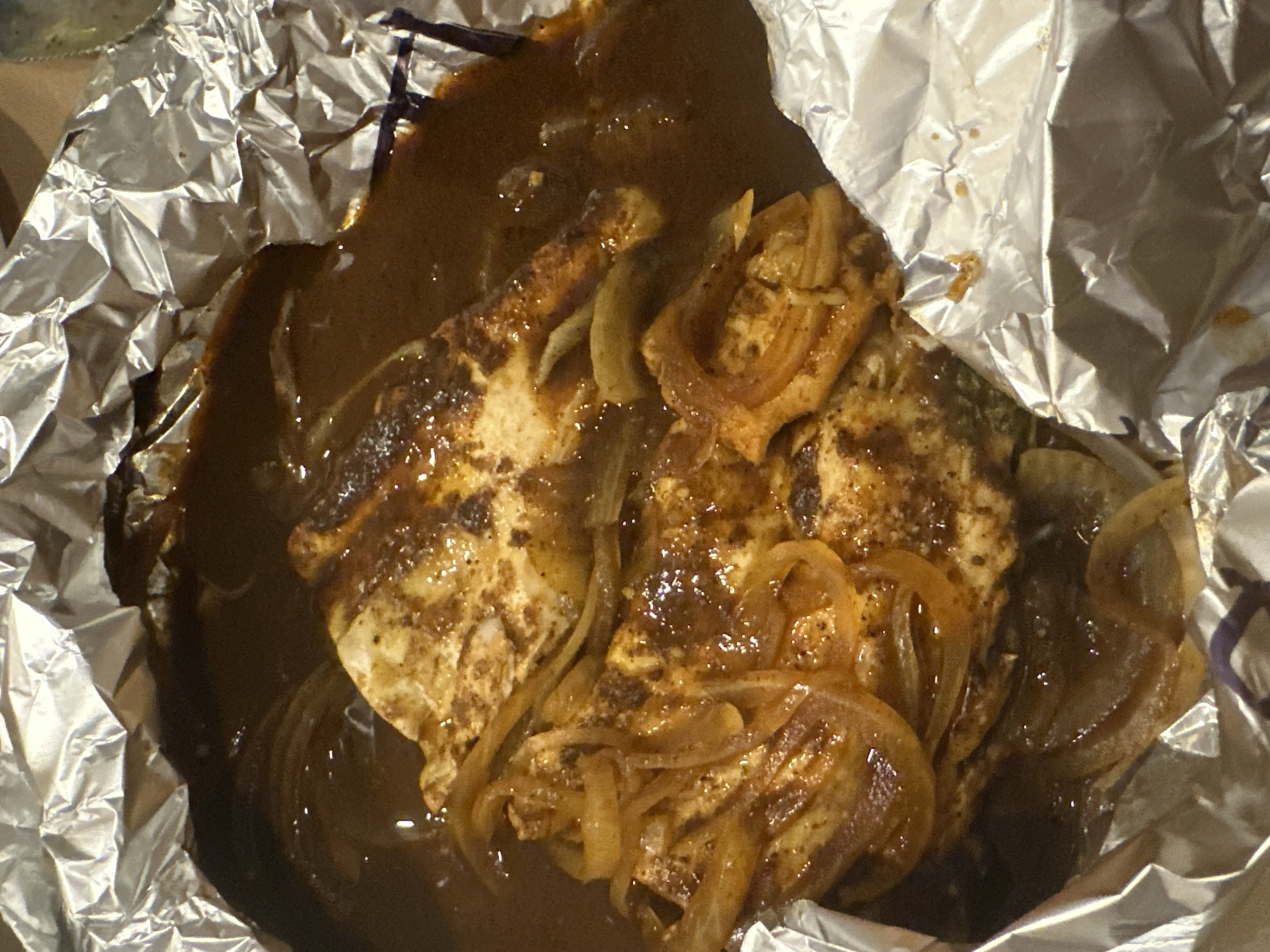 A close-up shot shows food wrapped in aluminum foil, likely a takeout container. The food appears to be slices of grilled chicken, covered in a dark brown sauce, with sauteed onions. The background is blurry and provides no clear context. The location in San Bernardino Tlaxcalancingo, Mexico, is not identifiable from the image.