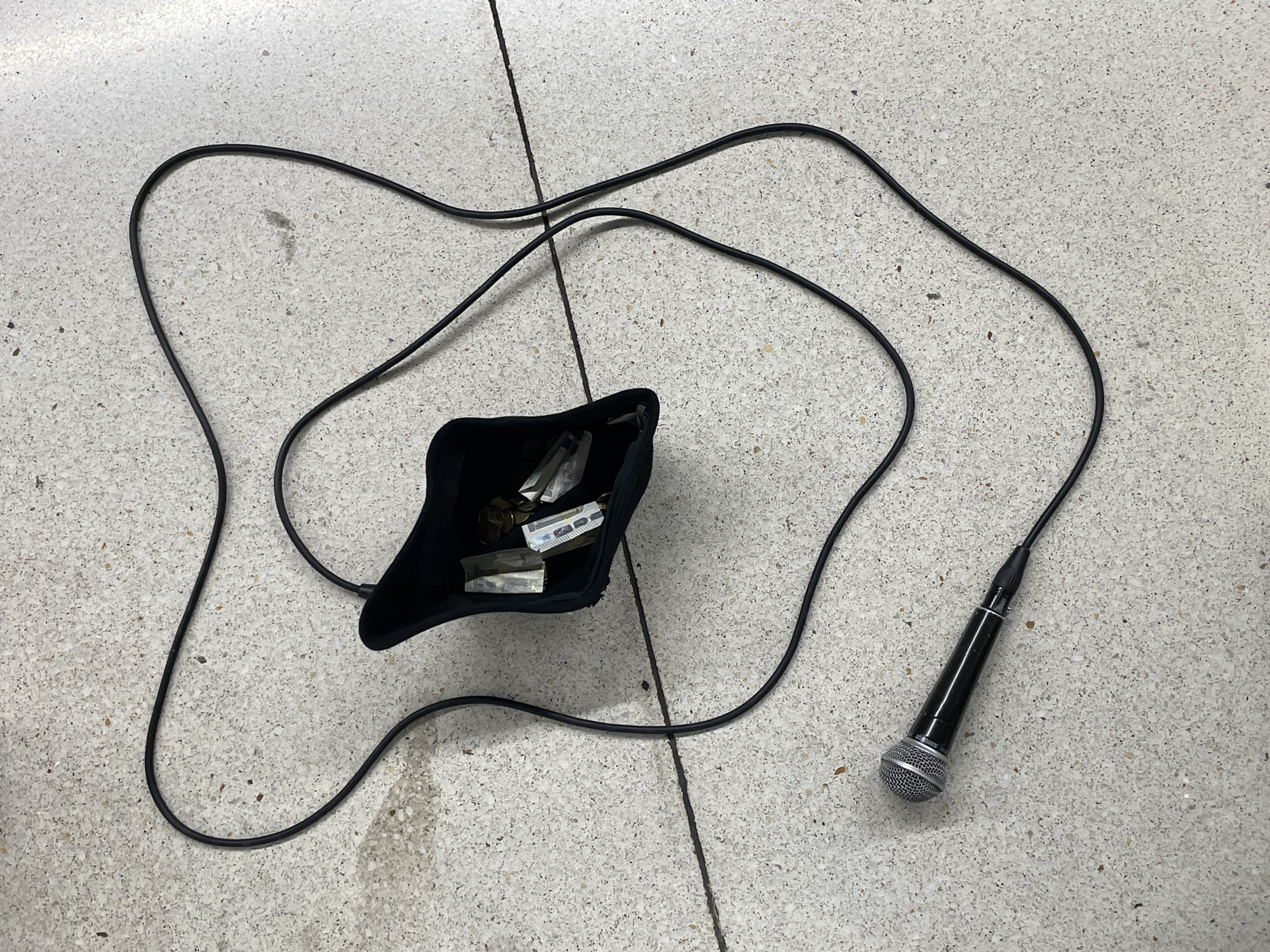 The image depicts a microphone attached to a long, black cable lying on a speckled, light-colored floor, possibly concrete. Next to the cable, a black cloth bag sits, containing what appears to be money and coins. There are no people present. The location in Barcelona, Spain is not apparent from the image.