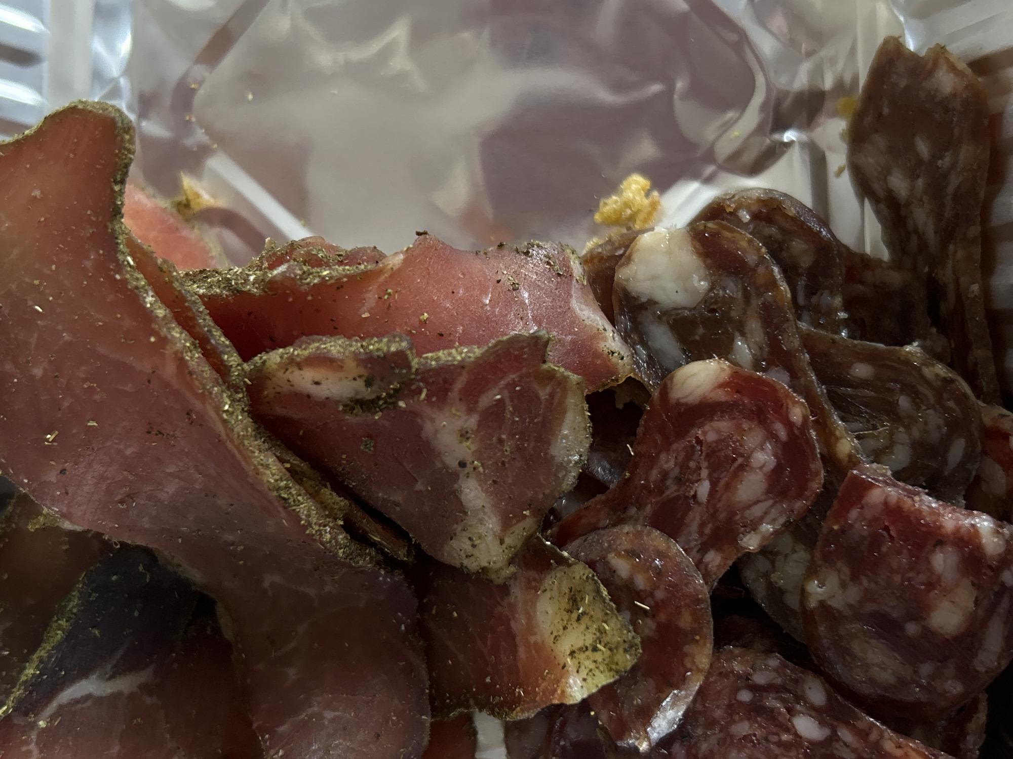 The image depicts a close-up view of cured meats inside a clear plastic container. On the left, there are slices of air-dried beef, possibly bresaola or similar, seasoned with herbs like pepper or dried parsley. To the right, there are circular slices of salami or sausage, displaying a mottled pattern of red meat and white fat. The container is partially visible at the top of the image. There's no indication of a location within Sofia, Bulgaria. No people, actions or interactions are apparent.
