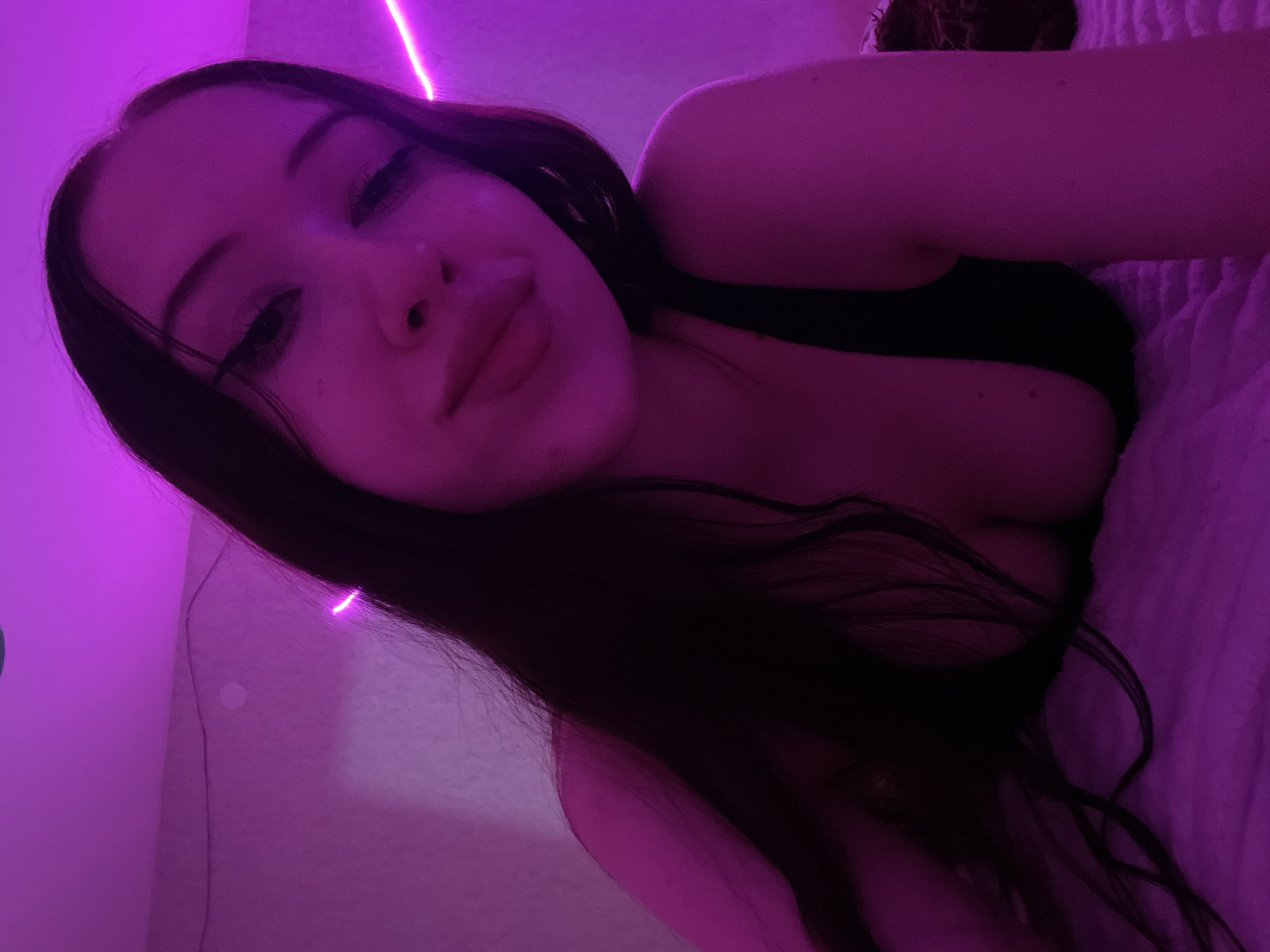 A young woman with long, dark hair is lying on a pink, fluffy surface, likely a bed or couch. She is wearing a black tank top. The scene is illuminated by vibrant pink LED lights, casting a strong purple hue over everything. The lighting creates a dramatic and intimate atmosphere. The background appears to be a wall, also bathed in pink light. The woman is looking directly at the camera, with her makeup, including winged eyeliner, clearly visible. Her pose suggests she is relaxed and comfortable in the dimly lit, colorful environment.