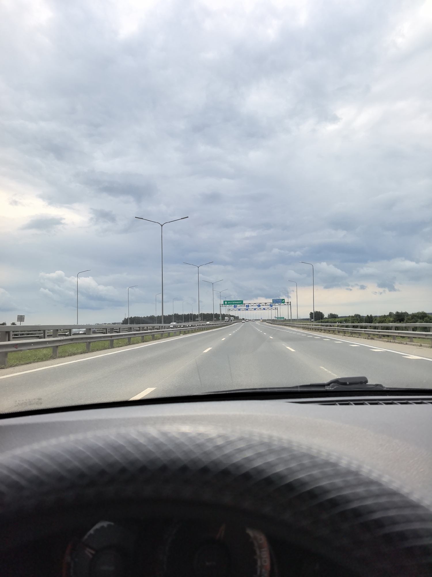 The image is taken from the driver's perspective inside a vehicle, looking forward along a multi-lane highway. The road is asphalt with white lane markings. On either side of the highway are metal guardrails and grassy shoulders, beyond which a line of trees is visible. Multiple streetlights are positioned along the road. Overhead signs indicate directions to "ЕКАТЕРИНБУРГ" (Yekaterinburg) and other destinations. The sky is overcast with heavy, grey clouds. The dashboard of the car is visible in the foreground, out of focus. This scene is consistent with a highway environment in the vicinity of Горный, Russia, likely a rural or suburban area given the presence of trees and open space alongside the road. There are no people visible in the image.