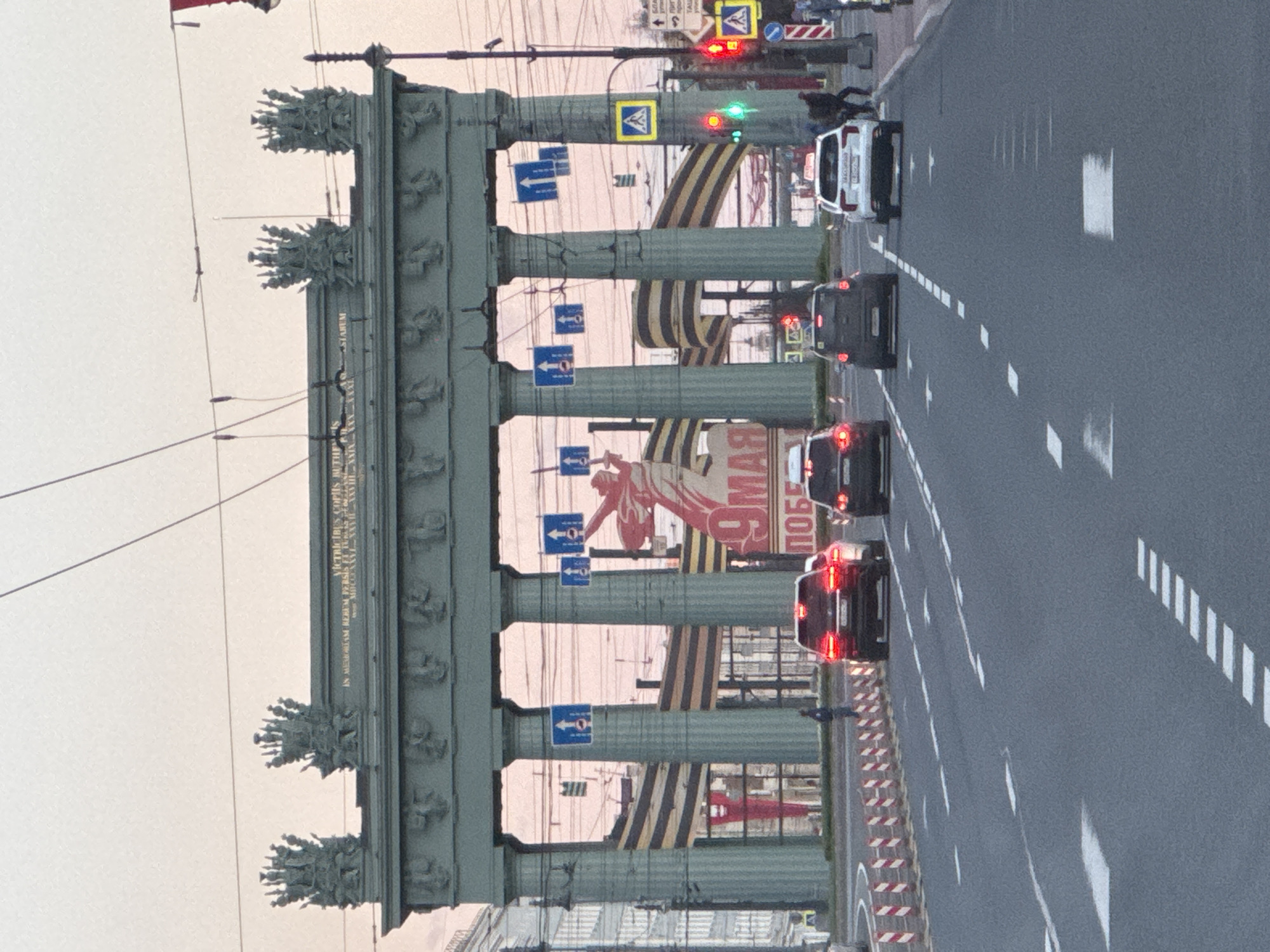 The image depicts a view through the Narva Triumphal Arch in Saint Petersburg, Russia. Cars are visible driving along the street through the archway. There are decorative elements visible on the archway, including columns, sculptures, and inscriptions. A large banner celebrating Victory Day, May 9th, is also displayed. Street signs and traffic lights are seen, and pedestrians are present near the street.