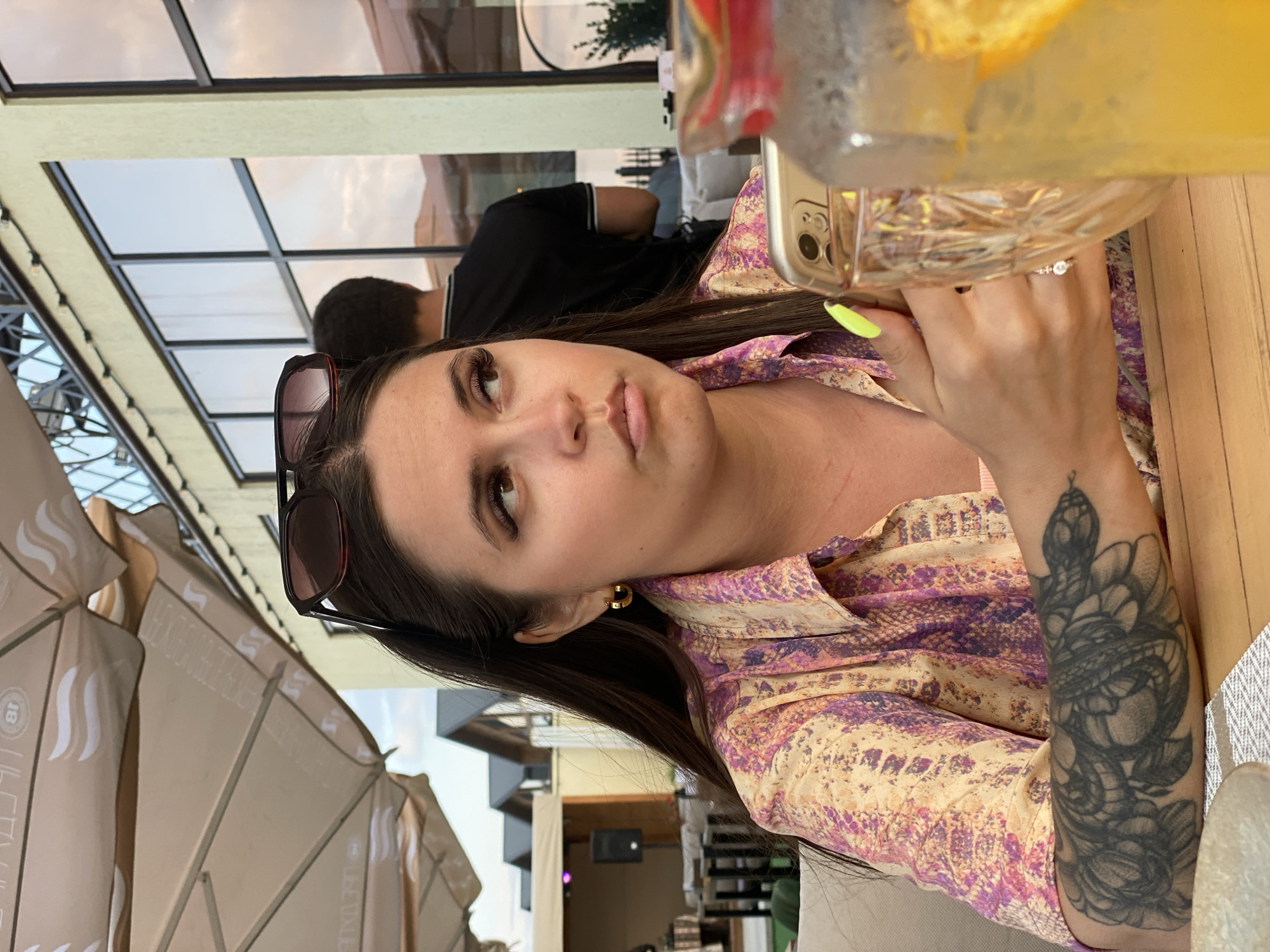 The image depicts a young woman sitting at an outdoor table, possibly at a restaurant in Derbent, Russia. She has long dark hair and is wearing a colorful, patterned shirt and gold hoop earrings. Sunglasses are perched on top of her head. She is looking to her right with a pensive expression. Her left arm, visible in the foreground, features a prominent tattoo of a snake. In her hands, she holds a smartphone and a glass pitcher filled with a yellow beverage and orange slices. A person in a black shirt is visible in the background, further back and out of focus. The environment appears to be a casual outdoor seating area, with umbrellas and buildings suggesting a developed urban setting. The lighting indicates it is daytime, possibly late afternoon, with soft, warm tones.