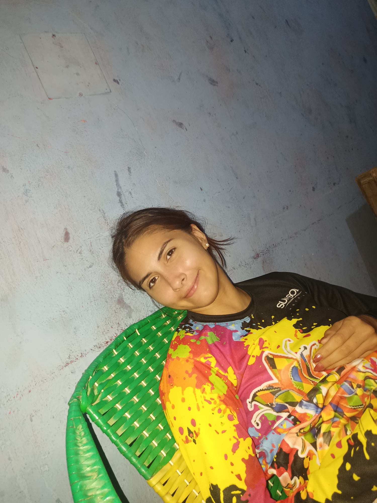 The image shows a woman sitting in a green and yellow wicker chair. She is wearing a black shirt with "Suiso Hotel" printed on it, covered partially by a brightly colored shirt or blanket with yellow, red, pink, and blue patterns. The background is a light blue wall with some markings or discoloration. The woman is smiling and looking at the camera. The location, Juan Griego, Venezuela, is not apparent from the image itself and would need to be determined through external information.