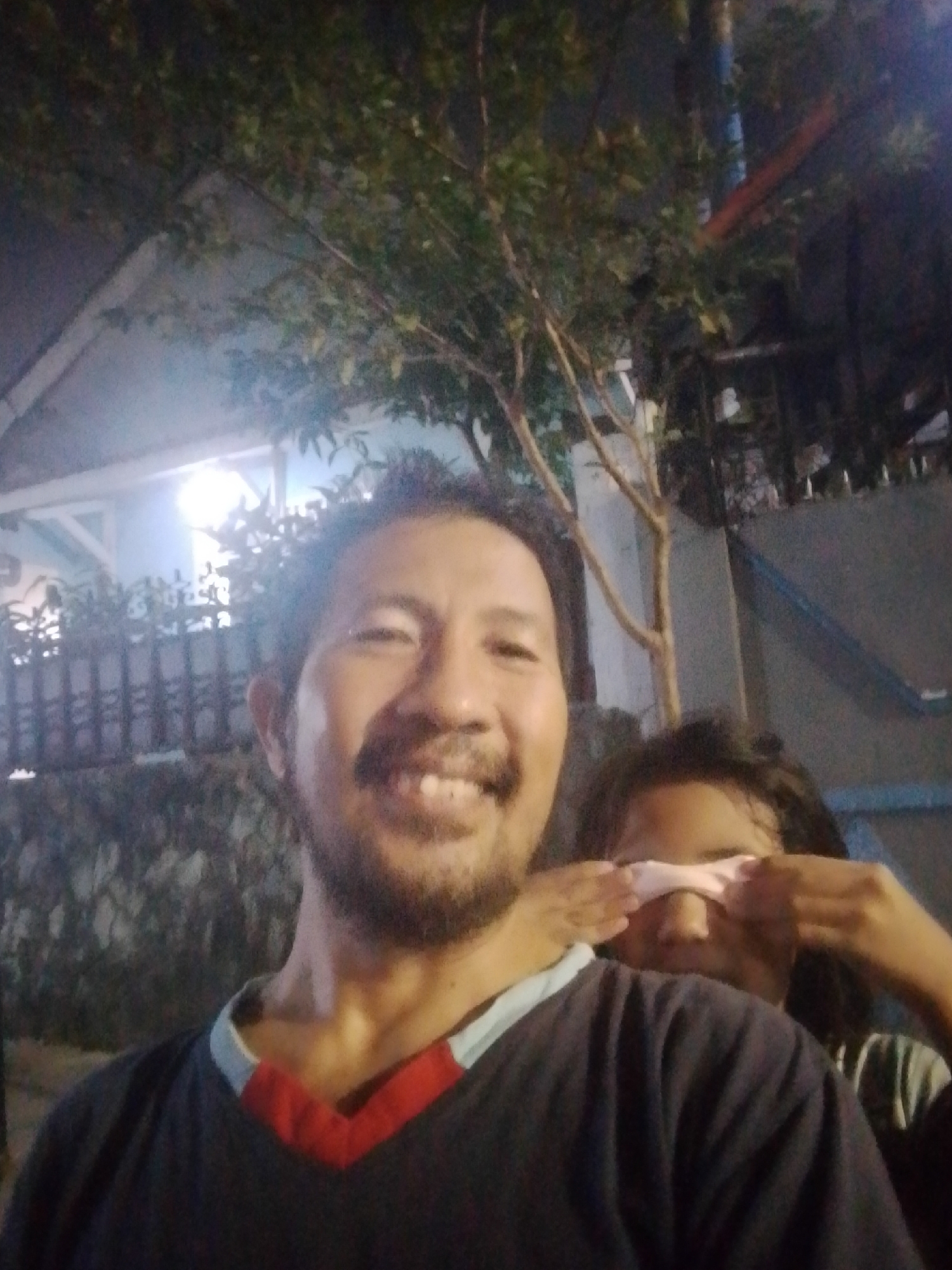 A selfie shows two people outdoors at night. A man in the foreground, facing the camera, smiles broadly. He has a beard and is wearing a dark v-neck shirt with red and white trim. Behind him, another person is partially visible, with their eyes covered by their hands. The background includes a tree with branches and leaves, a fence, and a building with lights. The scene appears to be in a residential area of East Jakarta, Indonesia.