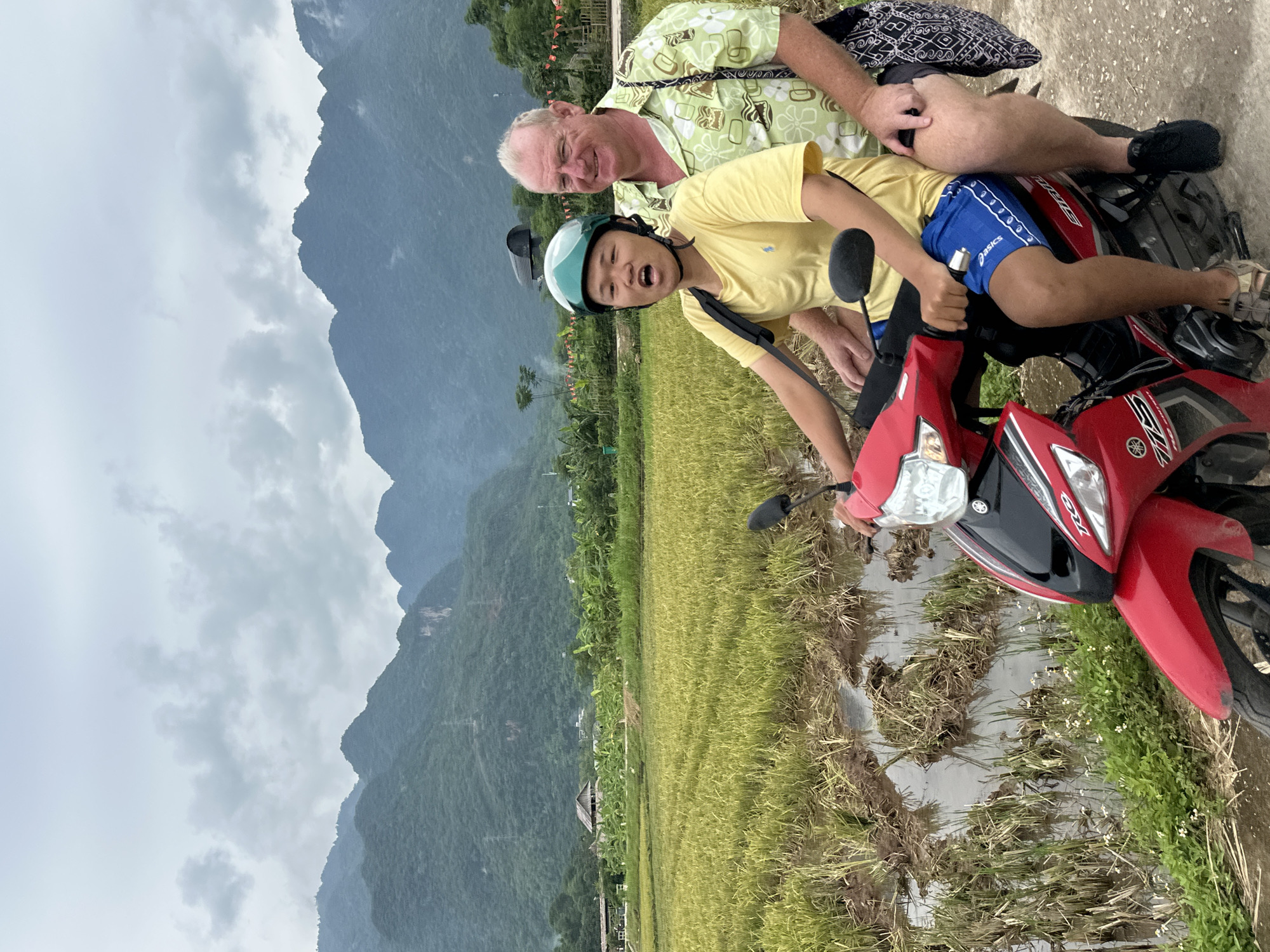 In Mai Chau, Vietnam, two individuals are depicted riding a red motorcycle on a dirt path beside a rice paddy. The person in front, wearing a green helmet, a yellow t-shirt, and blue shorts, appears to be a young male. He is leaning forward with his mouth open. The person behind him, a mature male with light skin and short hair, is wearing a light green patterned shirt and black shorts. He has his arm around the younger individual. The background features lush green rice fields, a cluster of banana trees, and mist-covered mountains under a cloudy sky. A small thatched-roof structure is visible in the middle ground. The overall scene suggests a rural and natural environment.
