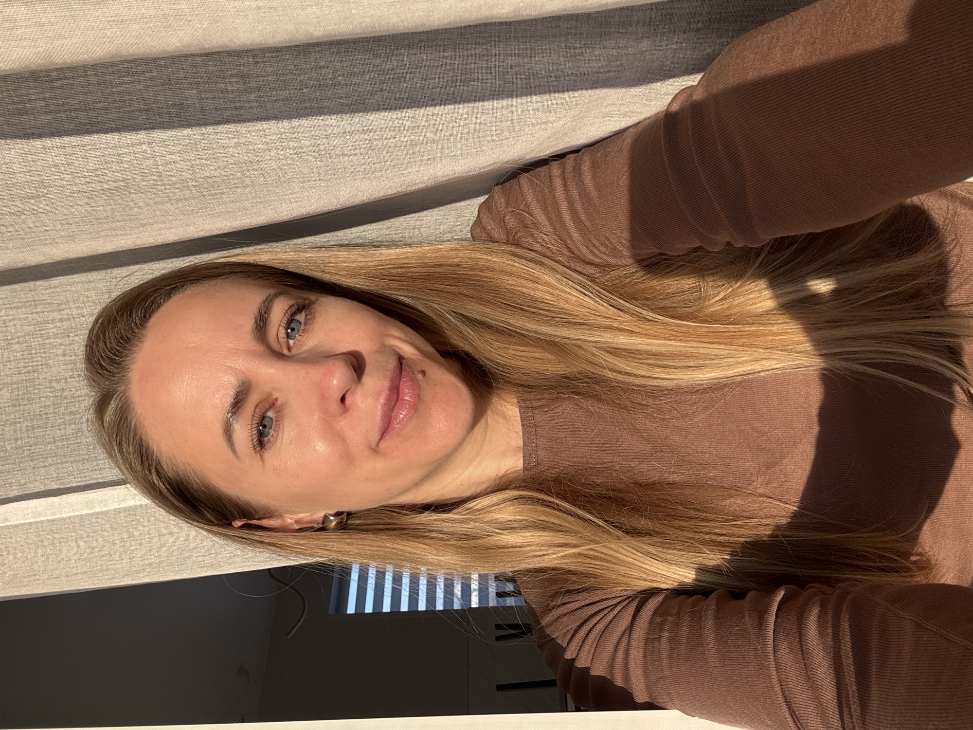 The image shows a woman with long blonde hair, fair skin, and blue eyes wearing a brown long-sleeved top. She is facing the camera with a slight smile. Behind her is a window with horizontal blinds, casting shadows across the wall and the woman's body. Based on EXIF data the image was taken on 2024:03:14 17:56:13 and geotagged at Chaikovsky, Russia (56°46'13.1"N 54°06'30.6"E). The background indicates an indoor setting, likely a residential space. The lighting suggests it is daytime.