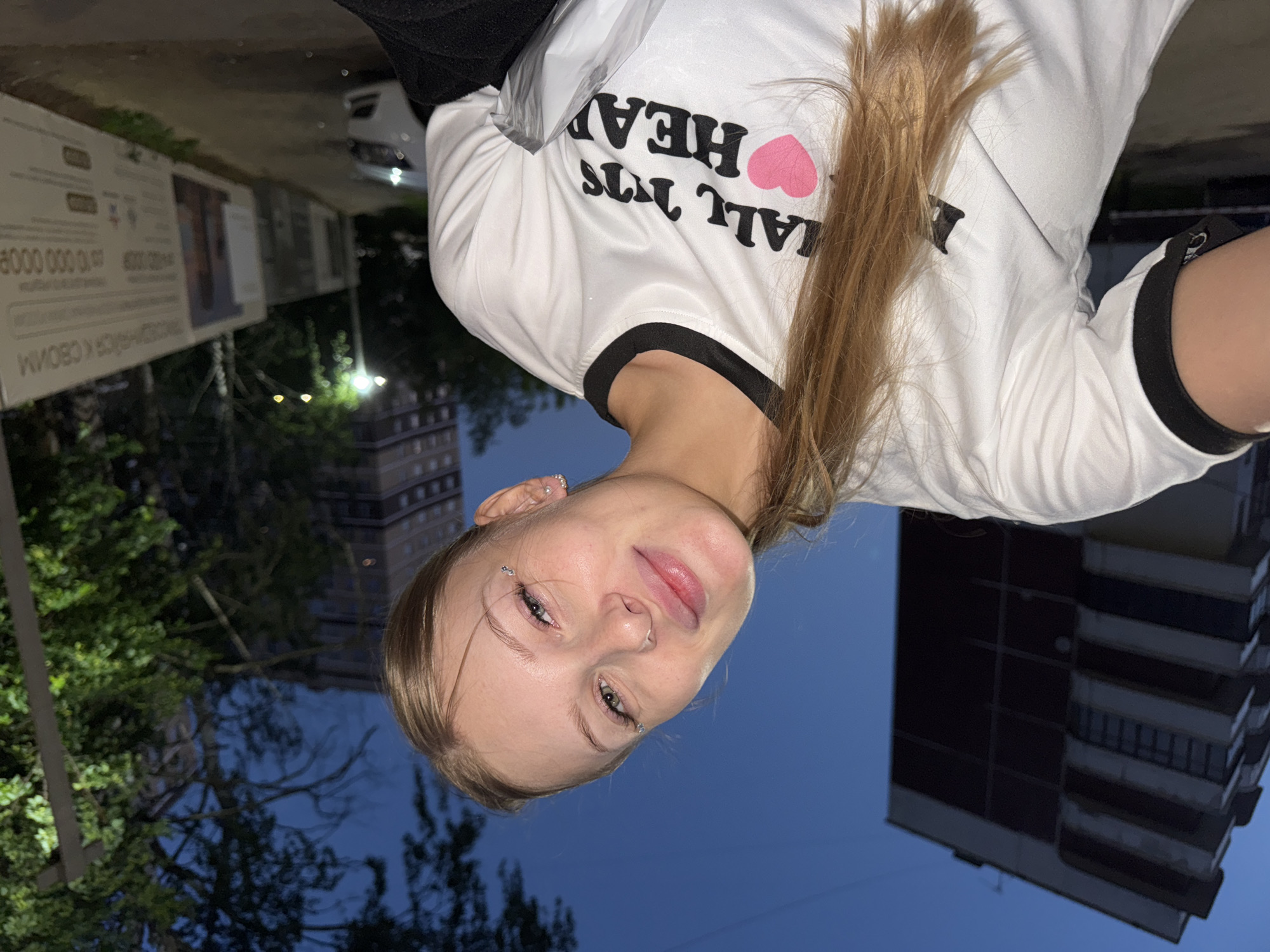 A young woman with blonde hair pulled back in a ponytail, wearing a white t-shirt with black trim and a graphic that says "ALL NUTS HEARTS", is taking a selfie outdoors at dusk. She has a nose ring and small silver embellishments on her face near her eyes. The background shows a tall, multi-story apartment building on the left, trees, and a streetlamp on the right. A white car is visible on the right side of the frame, parked near a construction site with large posters. The sky is a deep blue, indicating twilight. Based on the signage in the background, which appears to be in Russian and refers to a residential complex, the location is likely a new development area in the city of Perm, Russia.